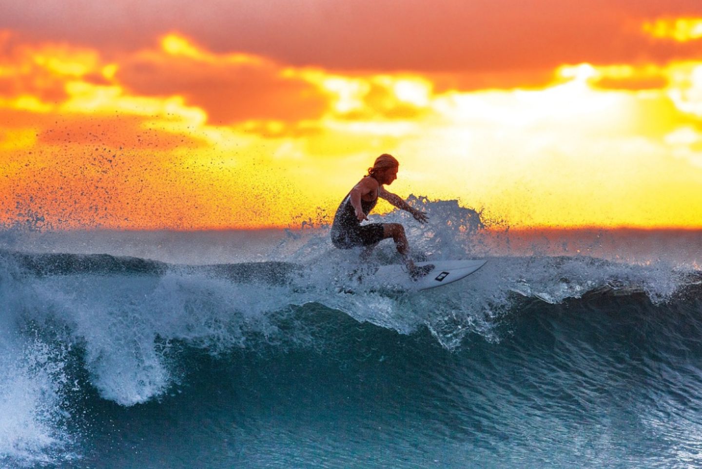 Large orange sunset behind a surfer about to catch a big wave.