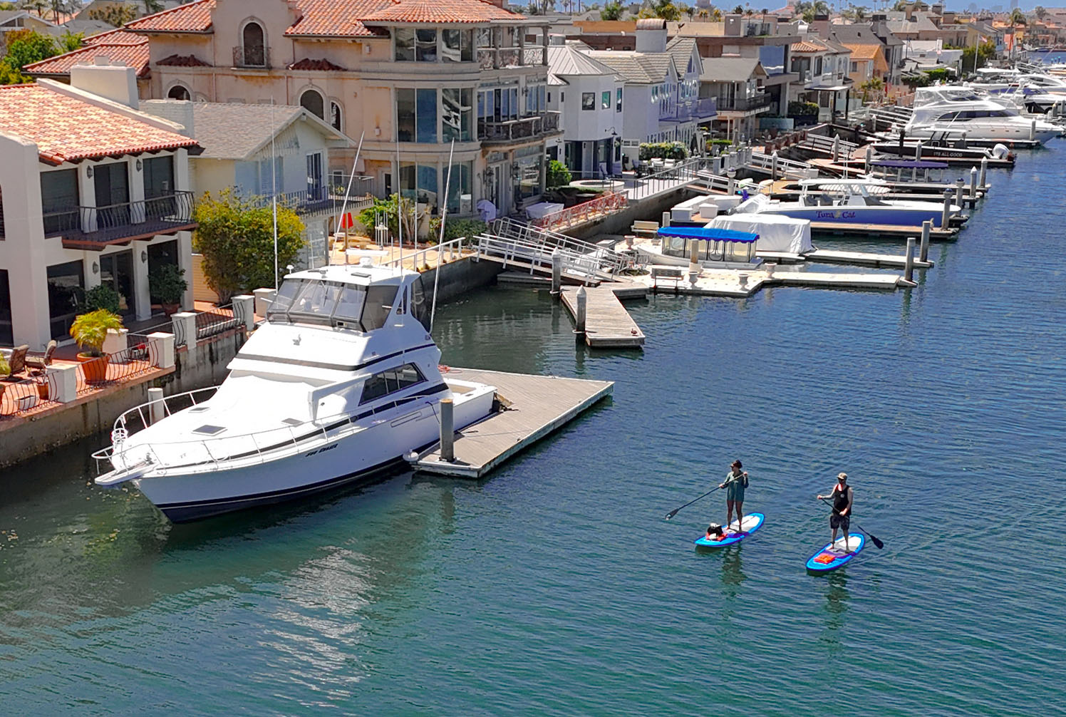 Aerial drone shot of two people paddle boarding through the canals of Huntington Harbour. Directly next to them is a yacht docked next to a row of homes. The water is calm and it is sunny.