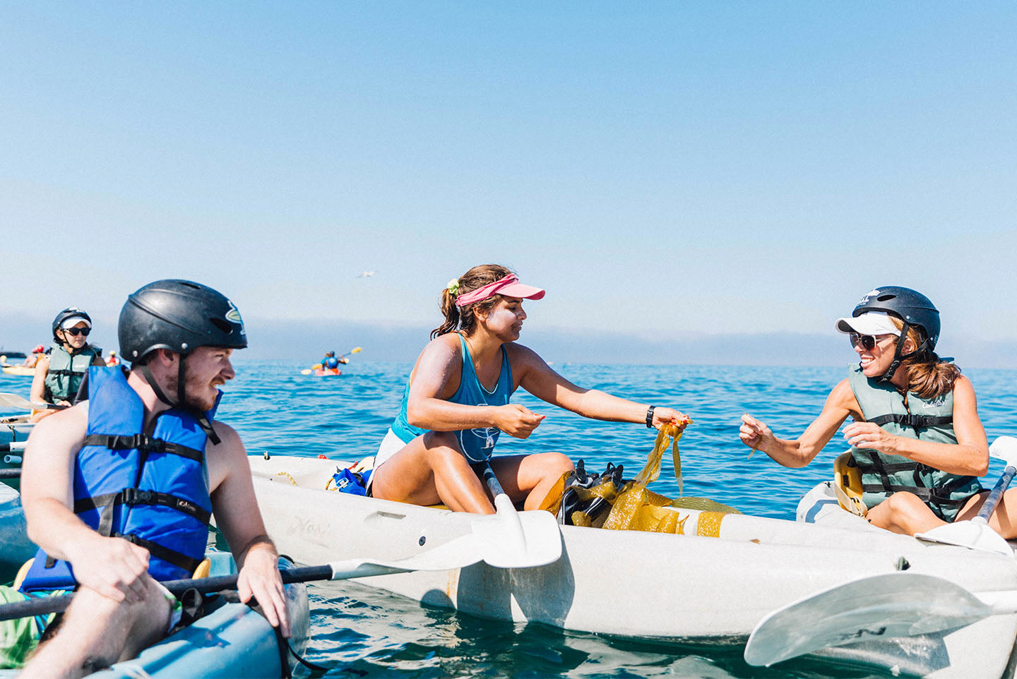 Three people sitting on kayaks in the ocean, passing a piece of kelp between each other.