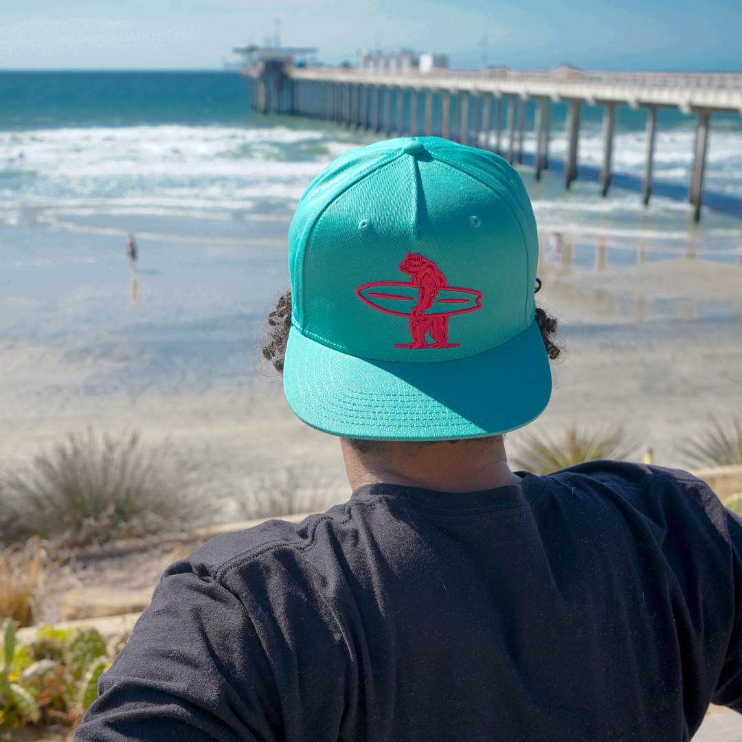 Corey posing with Brutus Connect hat worn backwards. Vibrant pink Brutus the bear contrasts against the aqua marine primary color of the cap.