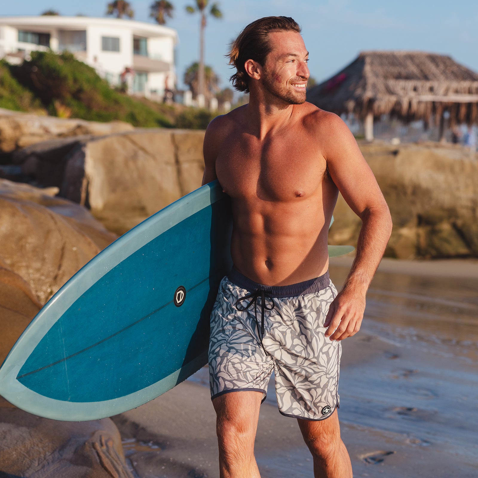 Man holding a surfboard walking along the beach wearing Everyday California boardshorts with leaf pattern and dark gray trim