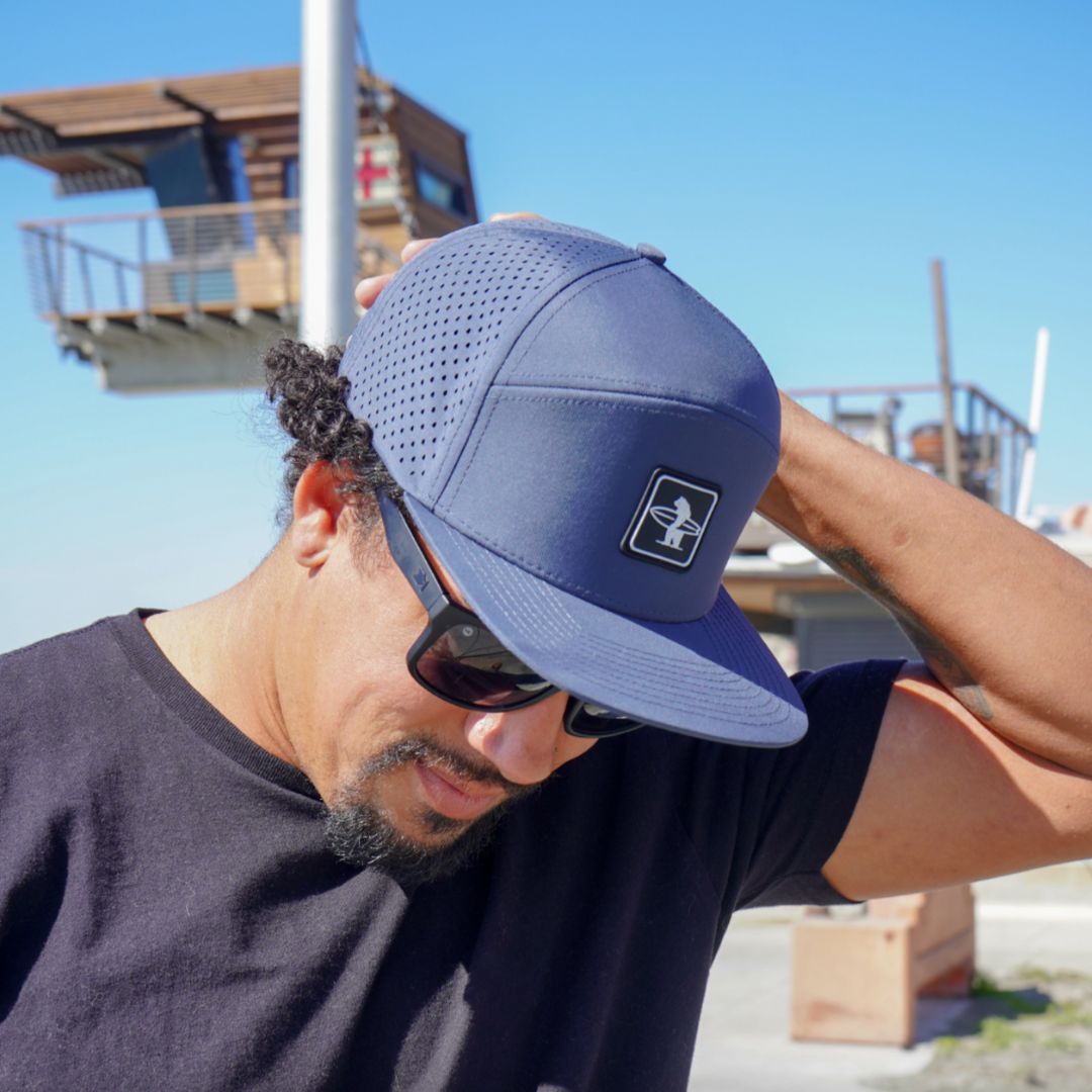 The sun shining on the Cormorant hat in navy in front of the beach lifeguard tower