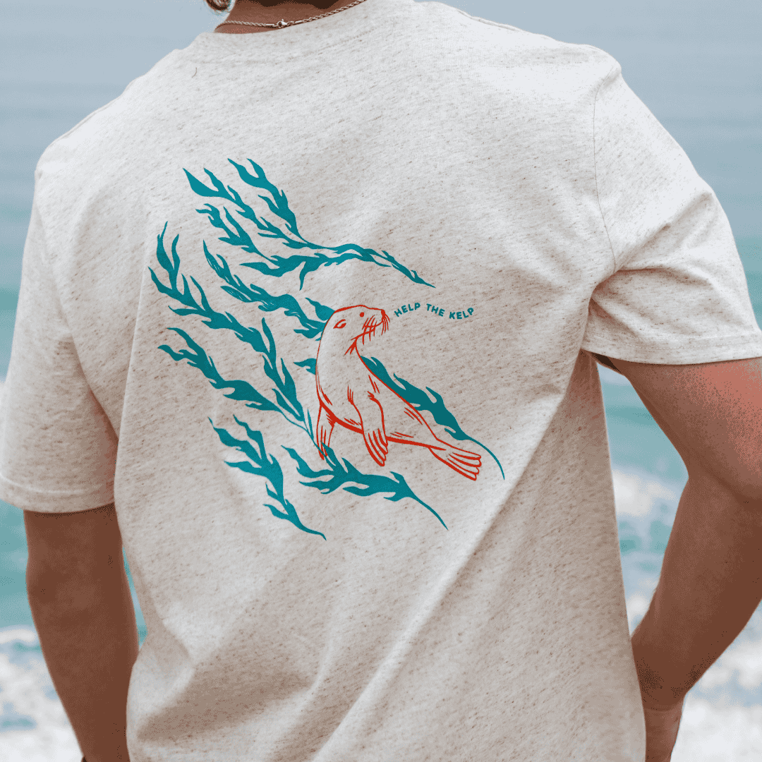 Person wearing a t-shirt with a sea lion and seaweed design on the back, with a blurred beach background.