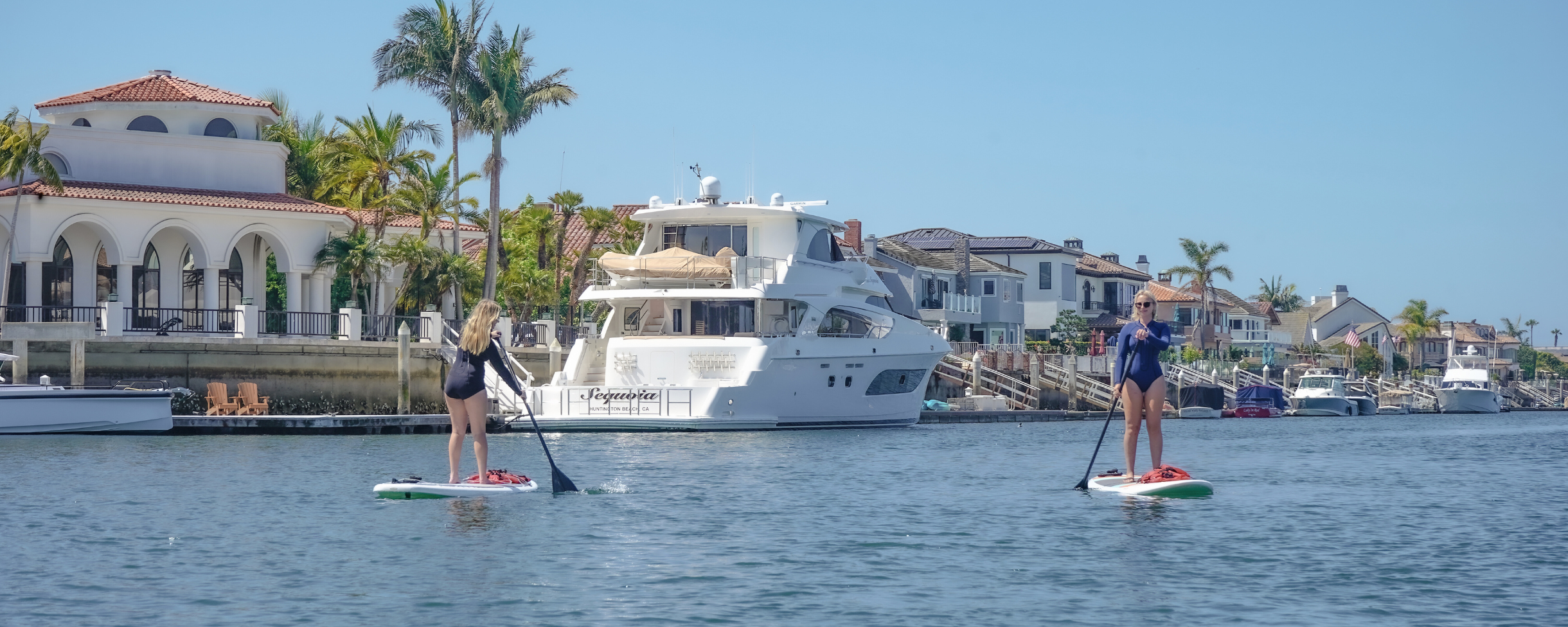 Two people paddleboarding on a calm Huntington Harbour with a marina and houses in the background.