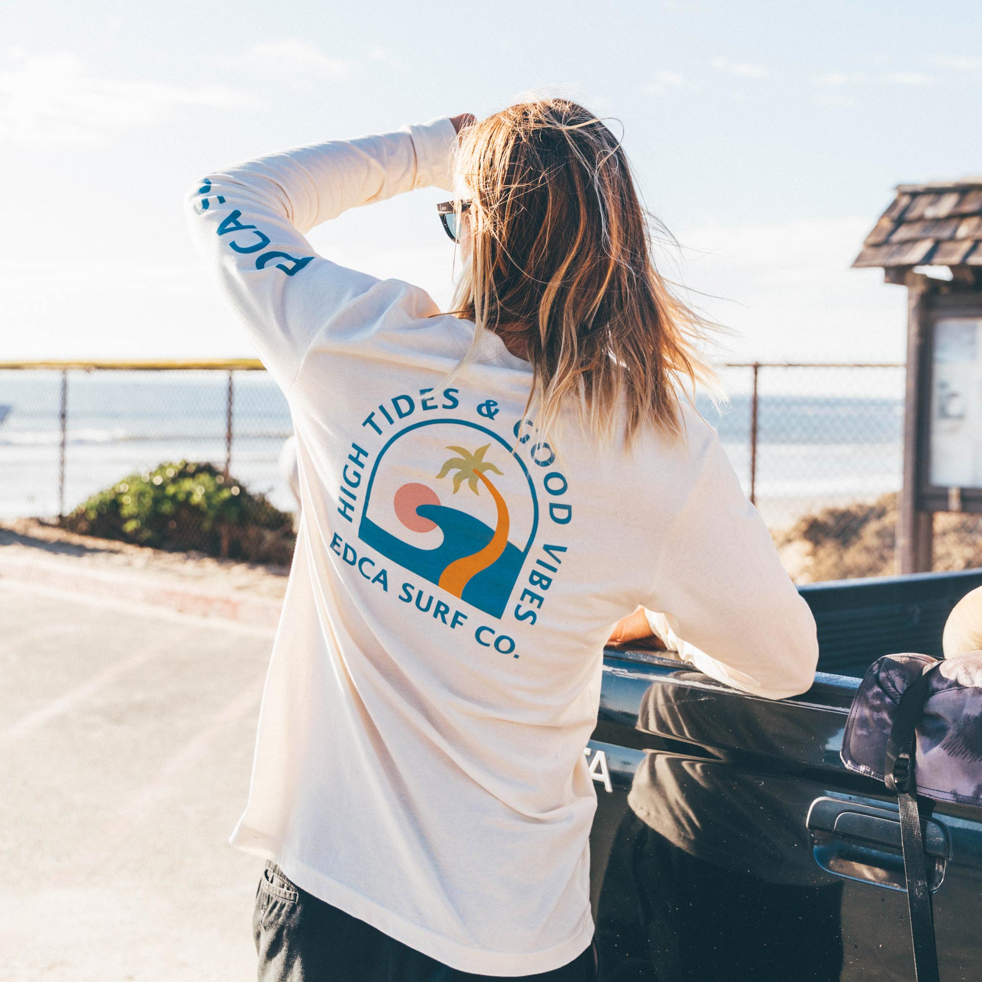 Large logo on the back with high tides and good vibes written on the back. Model is lifting left arm with EDCA Surf Co written down the side.