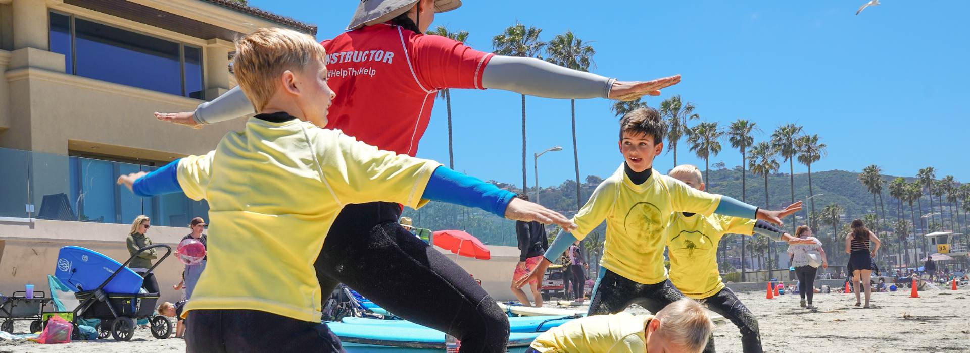 Children in yellow rashguards with a surf instructor on a beach with palm trees in the background during an Everyday California kid's summer surf camp in La Jolla.