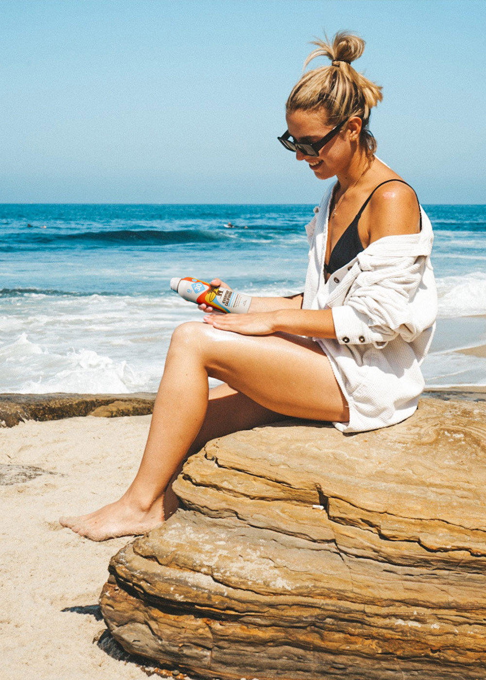 Photo of a girl sitting on a rock at the beach rubbing Everyday California Reef Safe Sunscreen into her skin.