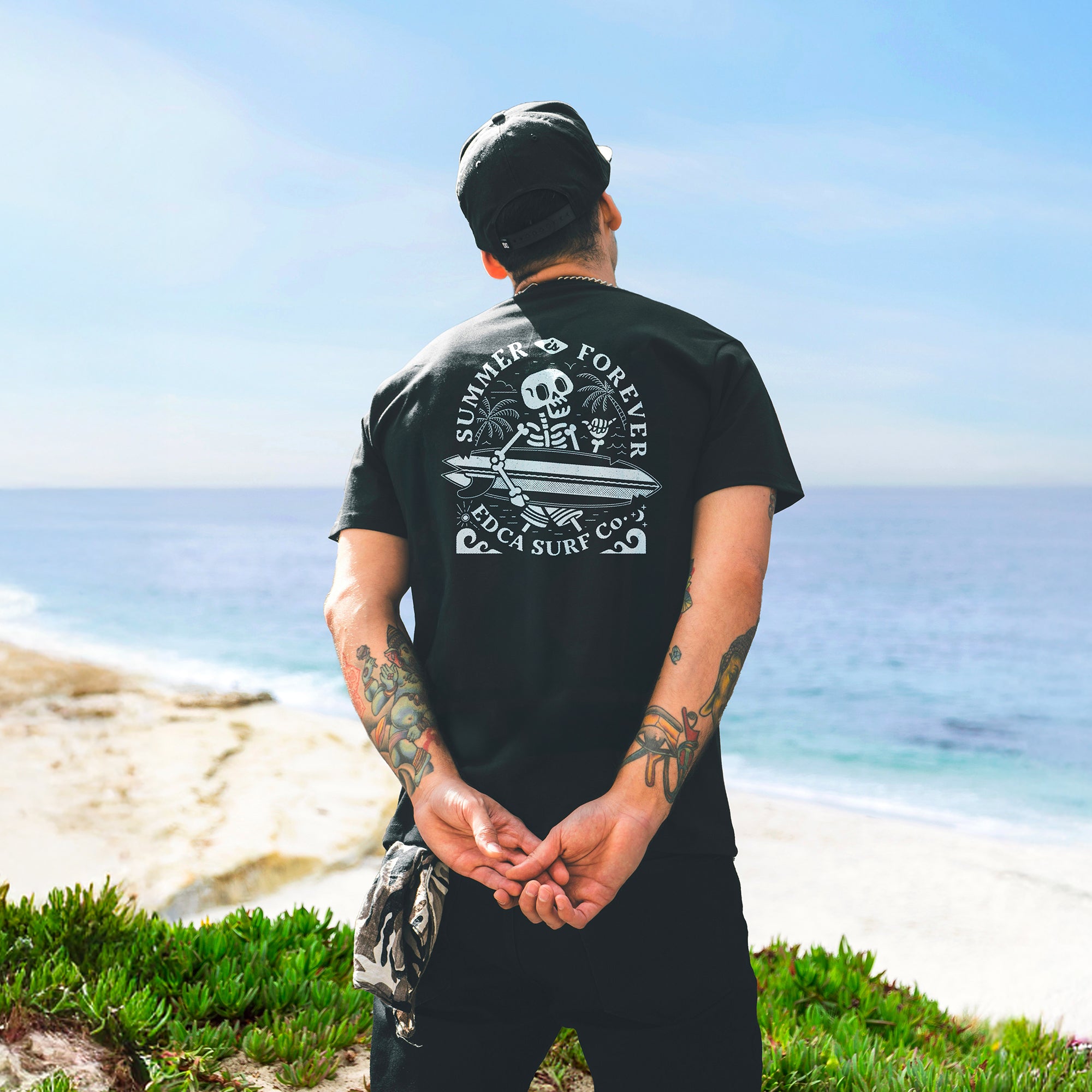 Man on the beach facing away from the camera, wearing a black t-shirt with a screen printed design of a skeleton holding a surfboard on it with the text Summer Is Forever above the image.