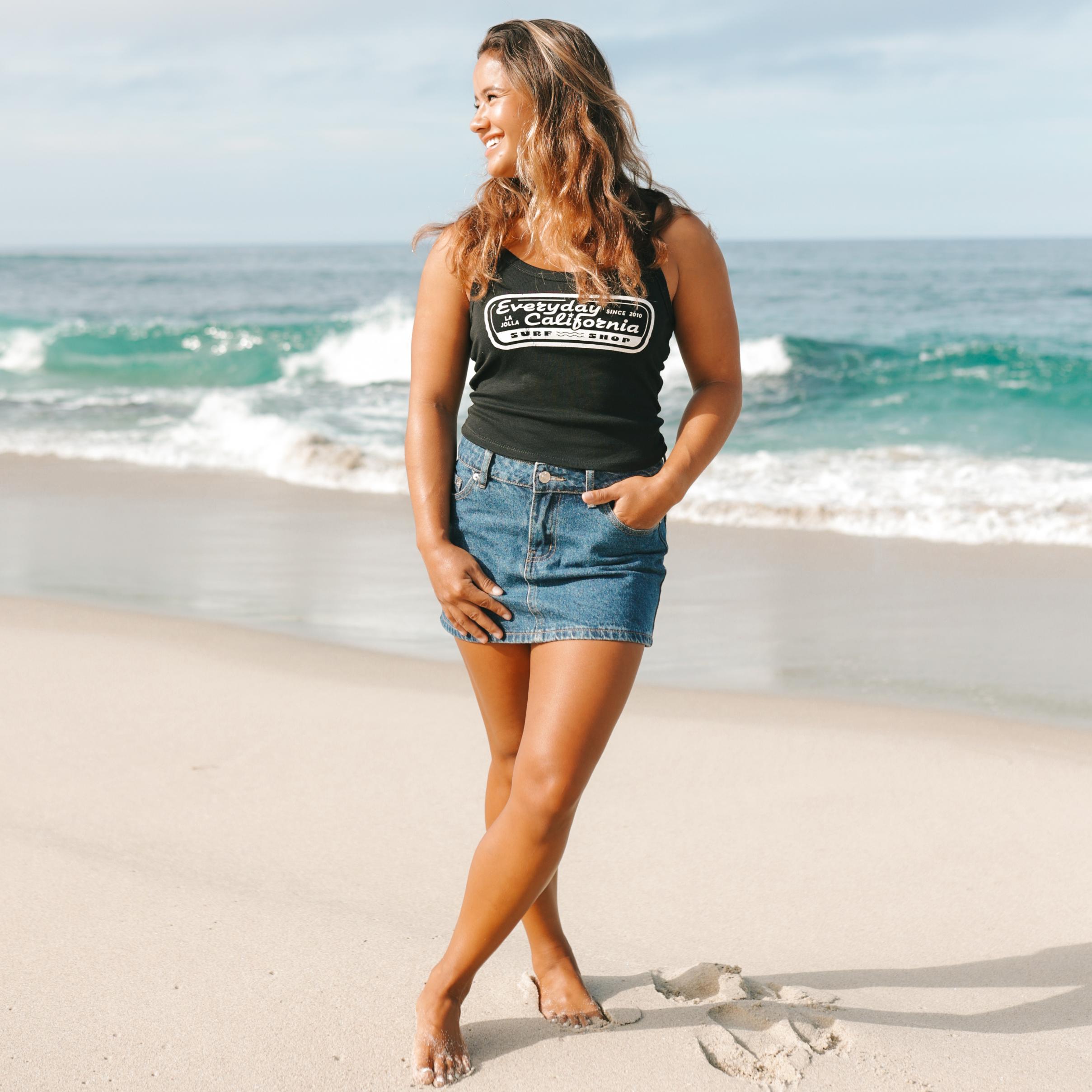Woman standing on a beach wearing a black tank top with Everyday California text and a denim skirt.