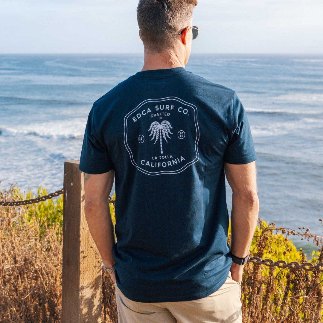 Man wearing a navy blue t-shirt with a palm tree design, standing by the ocean.