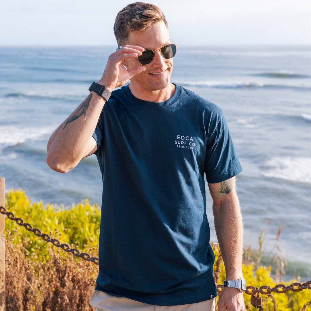 Man wearing a navy blue t-shirt with 'EDCA Surf Co' logo by the ocean.