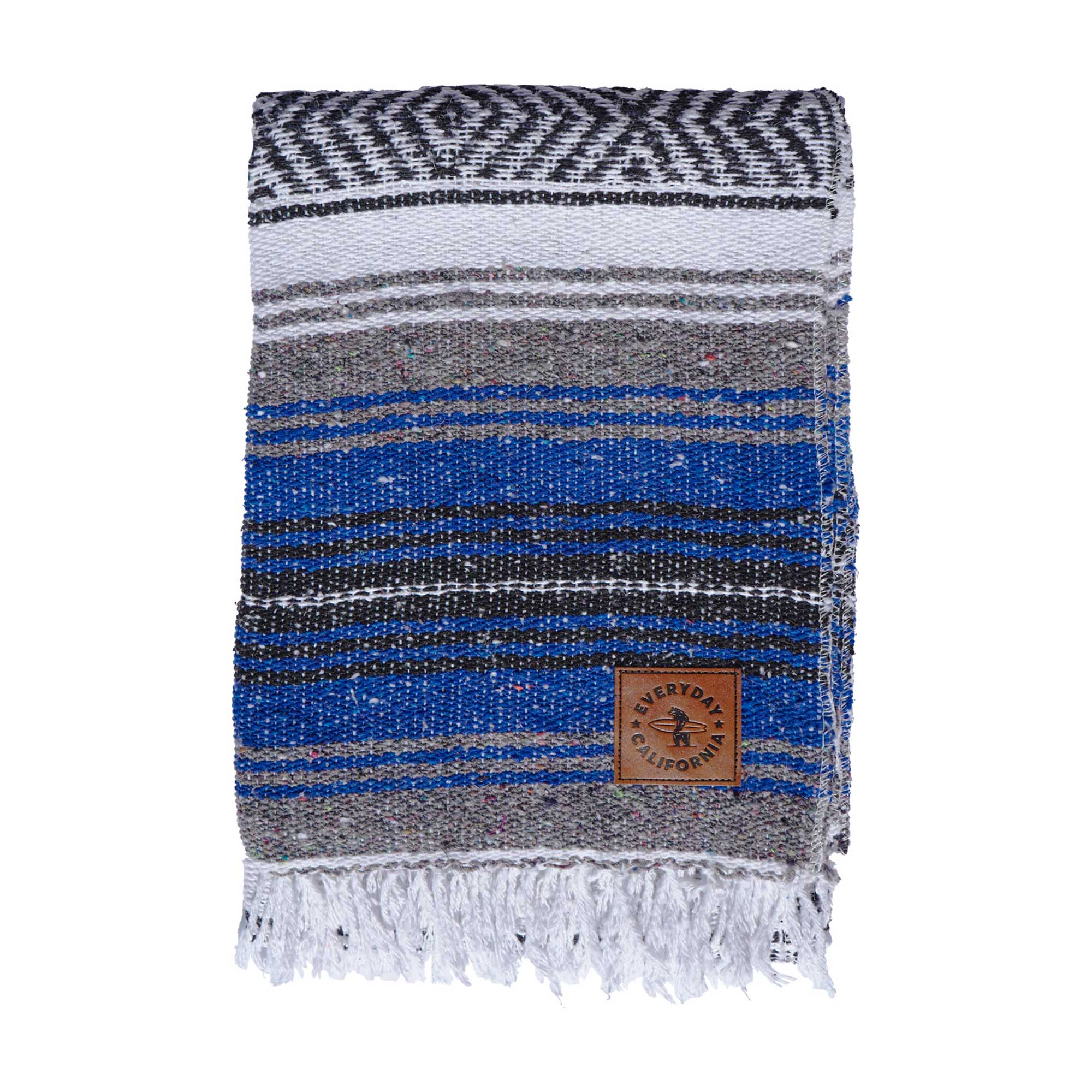A striped multicolor blanket with fringes, featuring shades of blue , gray, and white, with a vegan leather logo patch on the lower corner which features a bear holding a surfboard and the words 'EVERYDAY CALIFORNIA'.