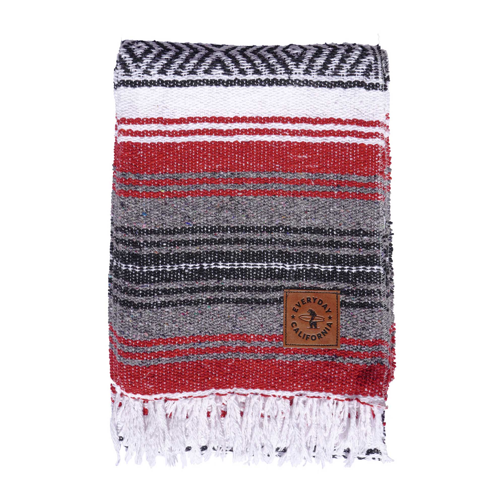 A striped multicolor blanket with fringes, featuring shades of red, gray, and white, with a vegan leather logo patch on the lower corner which features a bear holding a surfboard and the words 'EVERYDAY CALIFORNIA'.