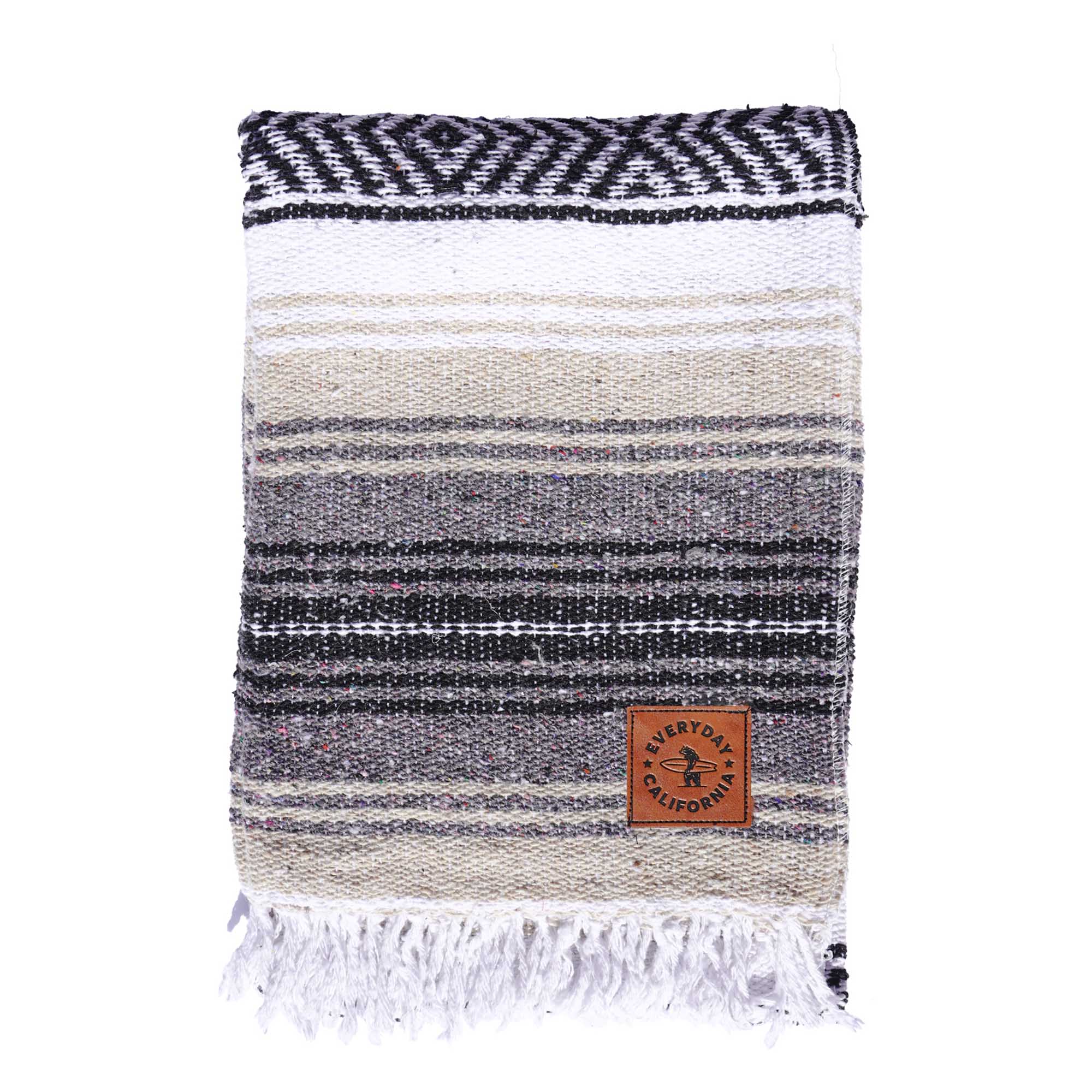 A striped multicolor blanket with fringes, featuring shades of gray and white, with a vegan leather logo patch on the lower corner which features a bear holding a surfboard and the words 'EVERYDAY CALIFORNIA'.