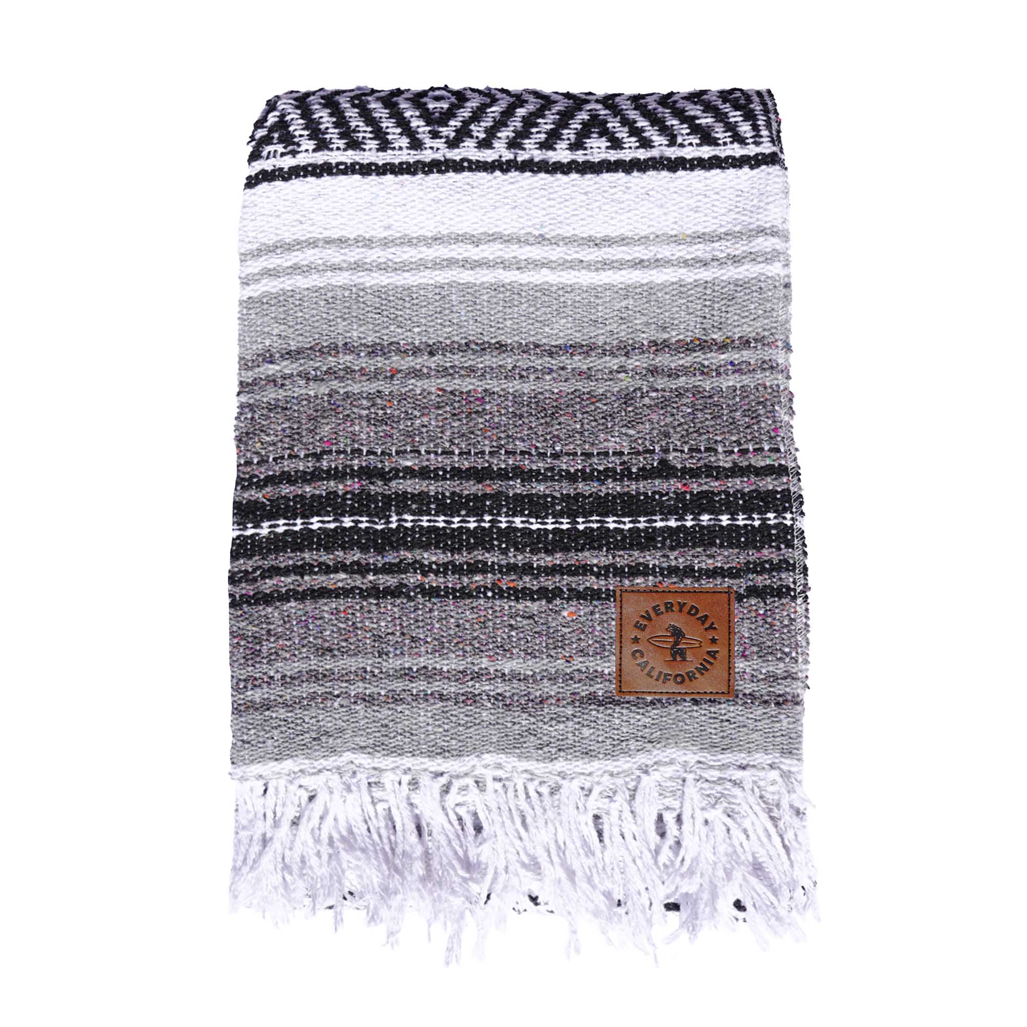 A striped multicolor blanket with fringes, featuring shades of gray and white, with a vegan leather logo patch on the lower corner which features a bear holding a surfboard and the words 'EVERYDAY CALIFORNIA'.