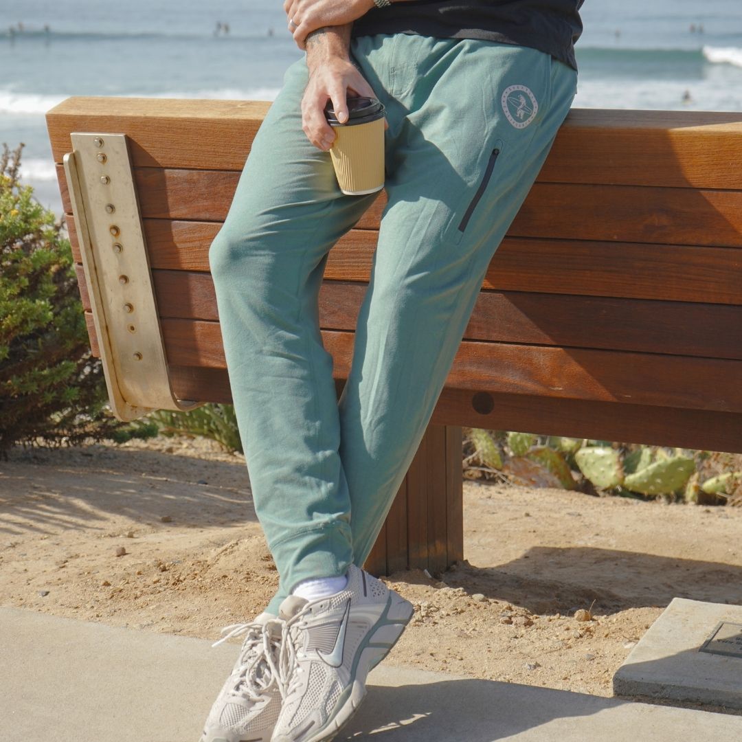 Man modeling the green Bayside Performance joggers featuring a small reflective vinyl logo near the hip pocket of a bear holding a surfboard.