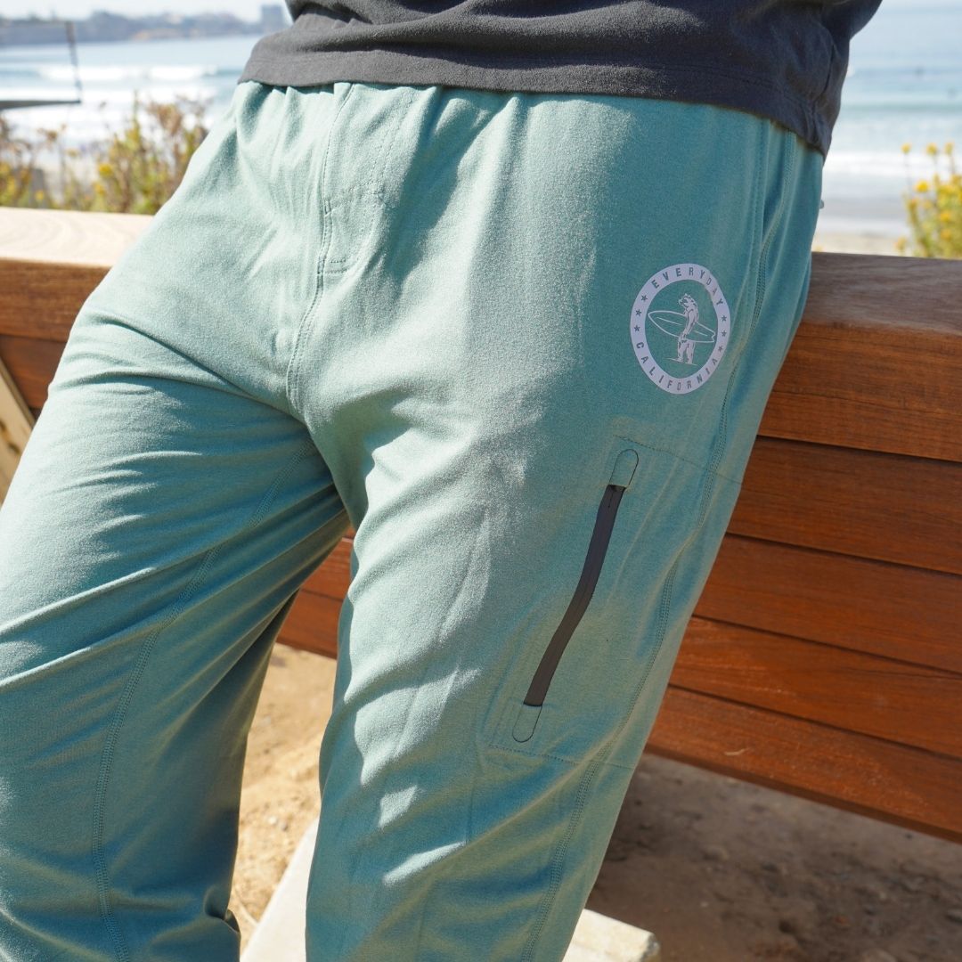 Man modeling the green Bayside Performance joggers featuring a small reflective vinyl logo near the hip pocket of a bear holding a surfboard.