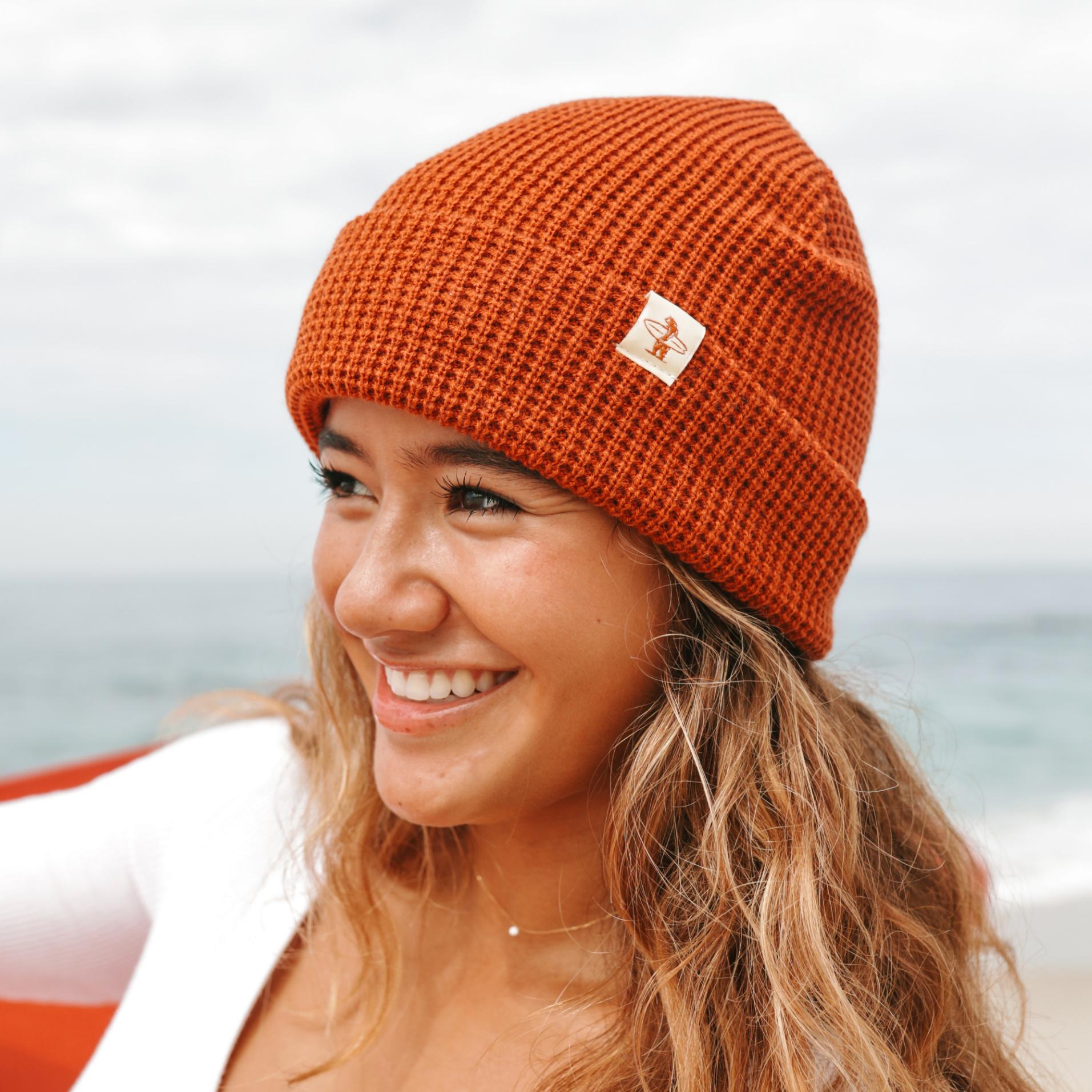 Woman wearing an orange knit beanie with a logo, smiling outdoors.