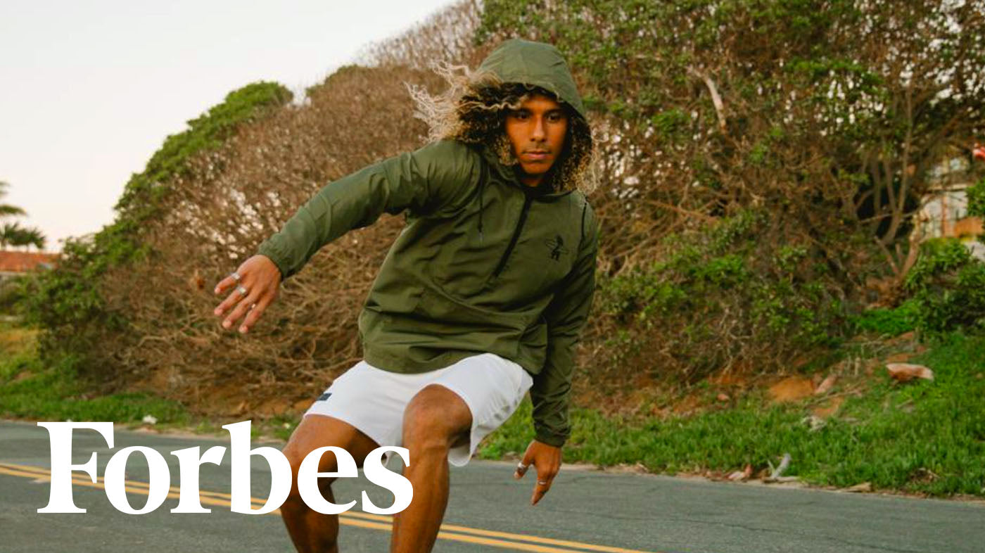 Photo of a man skateboarding down the middle of the street with a cliff behind him, he is wearing a green windbreaker jacket and white shorts