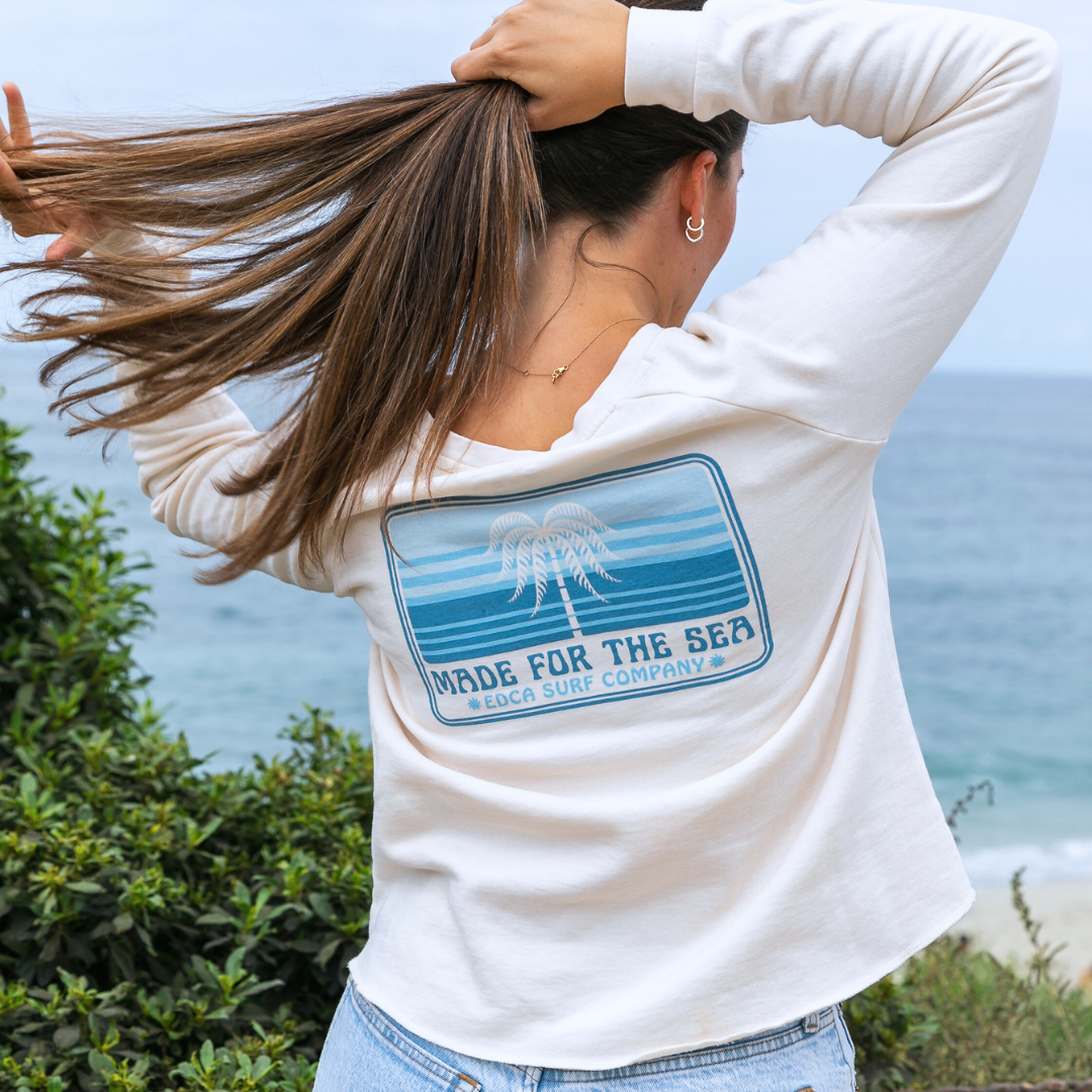 Back view of female model wearing a white Everyday California crewneck. The logo reads "Make for the Sea. EDCA Surf Company"