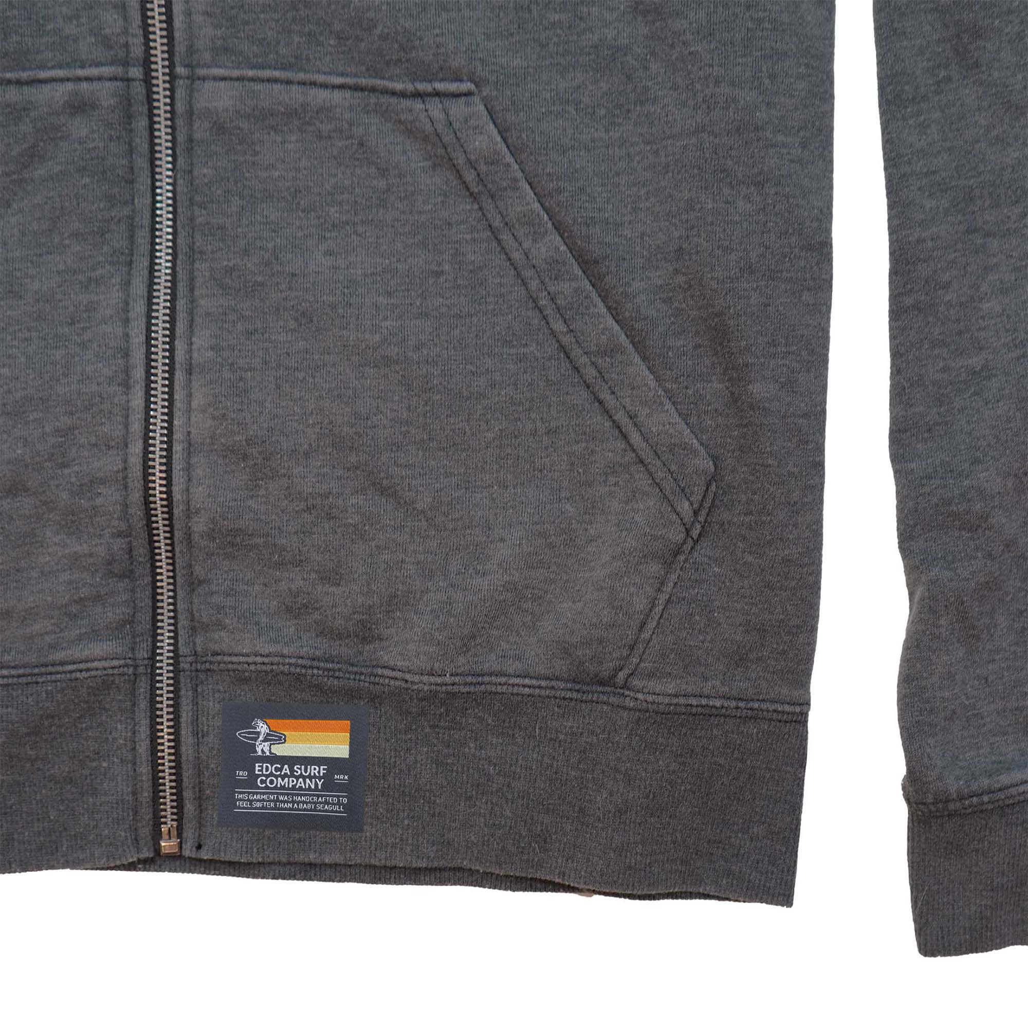 Closeup image of the woven label on the mavericks hoodie, which sits on the bottom rib of the hoodie near the zipper. The label has a dark gray background, and a bear holding a surfboard on it with the words "EDCA SURF COMPANY" in large text.