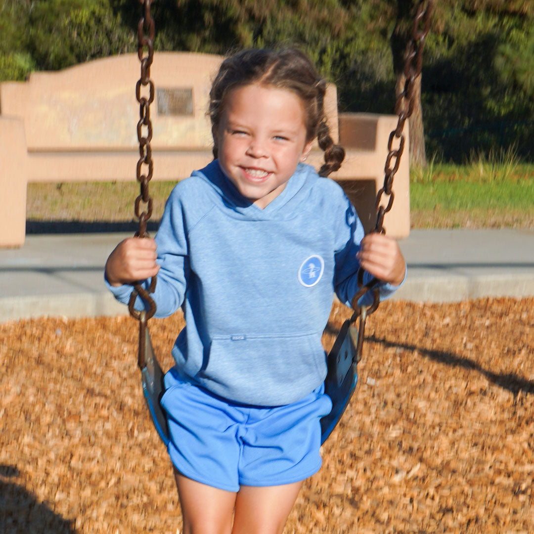 A young girl smiling while sitting on a swing at a playground, wearing a heathered blue hoodie with a small white circular logo on the chest and matching blue shorts.