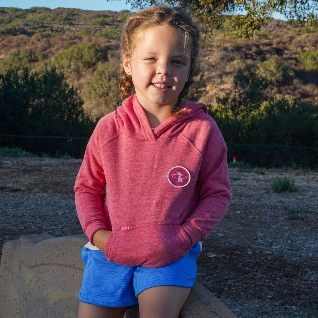 A young girl smiling outdoors while wearing a heathered pink hooded sweatshirt with a circular logo on the left chest and bright blue shorts, seated on a stone wall with hills and trees in the background.
