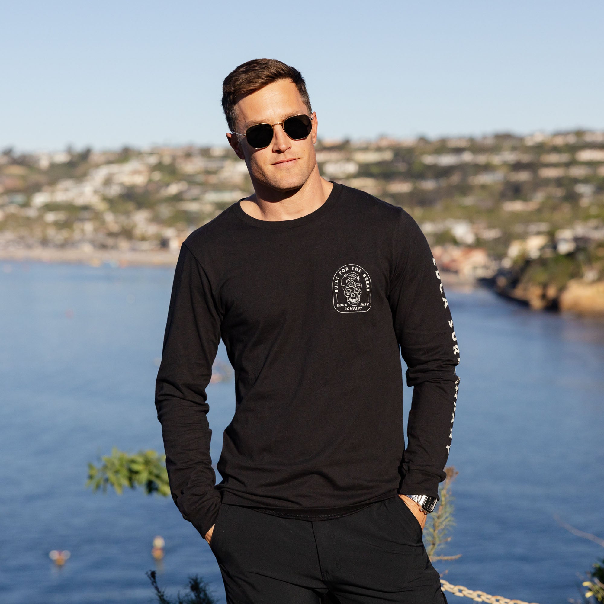 Man wearing a black long-sleeve shirt with a logo, standing by a waterfront with a scenic background.