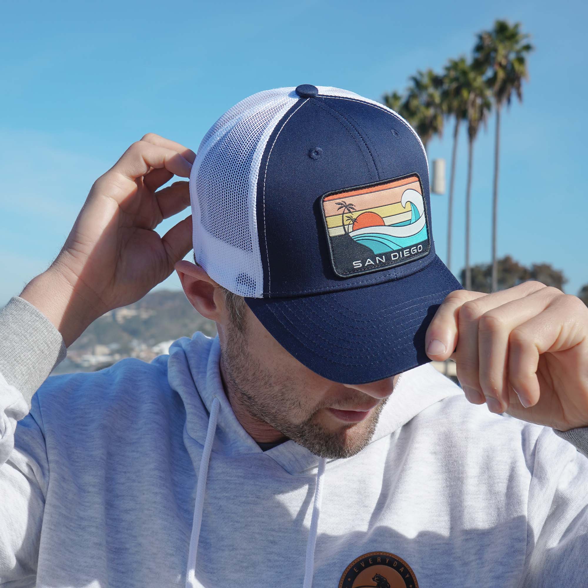 Man modeling a navy blue snapback with white mesh featuring a patch of an ocean wave, the sun, a palm tree, and the words 'San Diego California' on it.