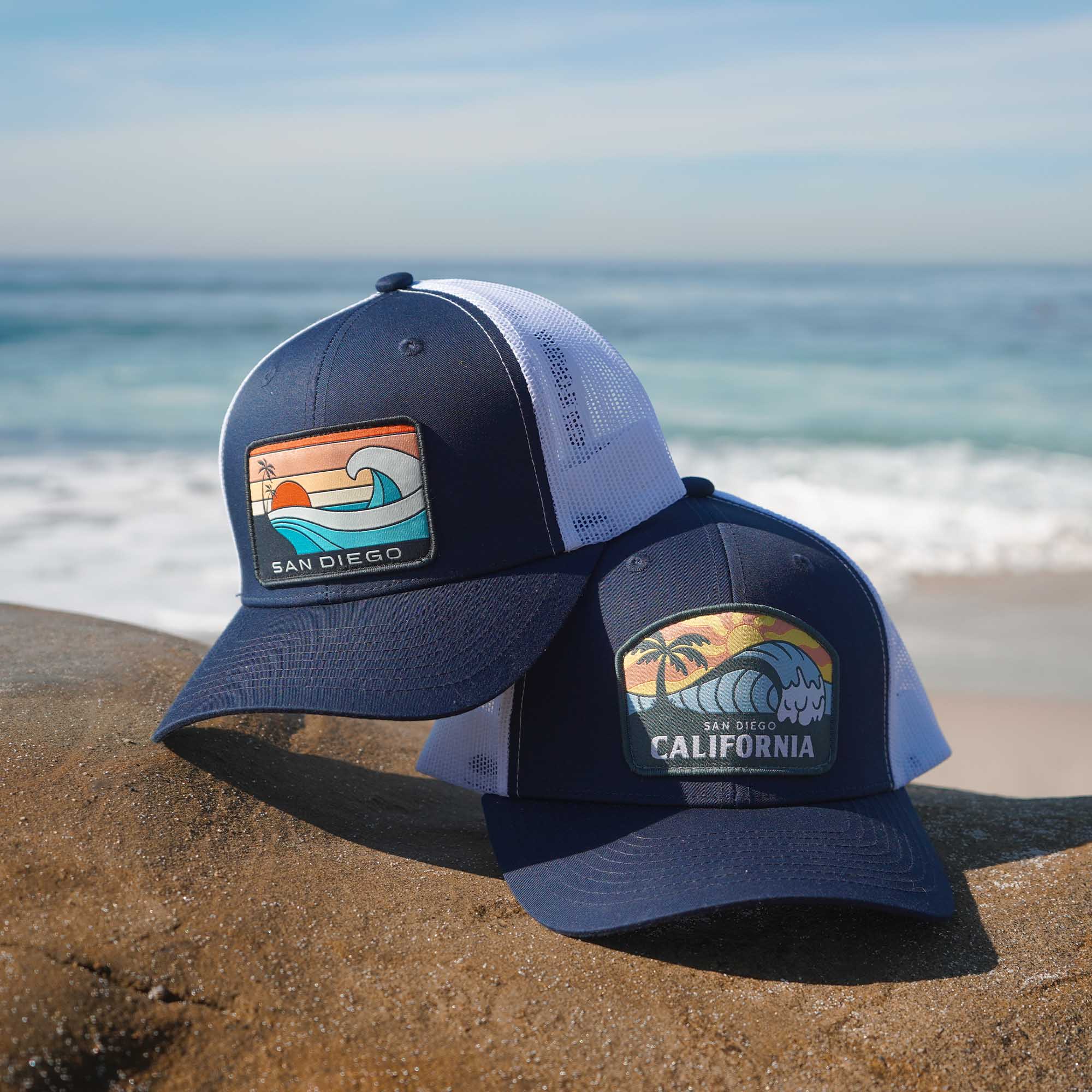Navy blue snapback with white mesh featuring a patch of an ocean wave, the sun, a palm tree, and the words 'San Diego California' on it, and a different navy blue snapback with white mesh featuring a patch with ocean waves, the sun, two palm trees, and the words 'San Diego' on it.