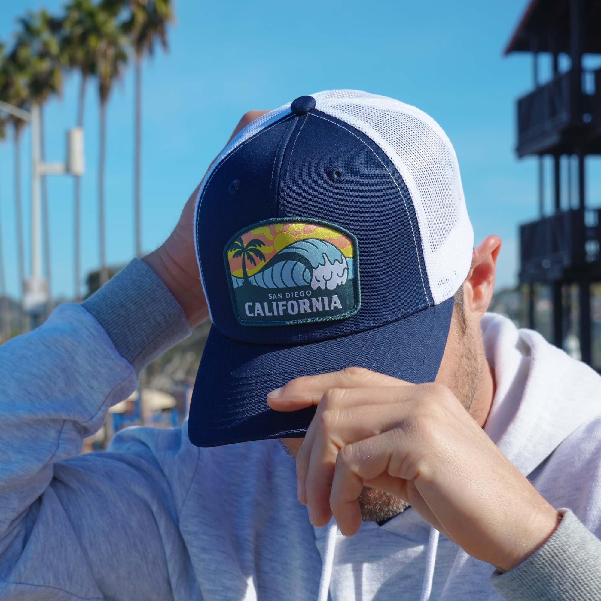 Man modeling a navy blue snapback with white mesh featuring a patch of an ocean wave, the sun, a palm tree, and the words 'San Diego California' on it.