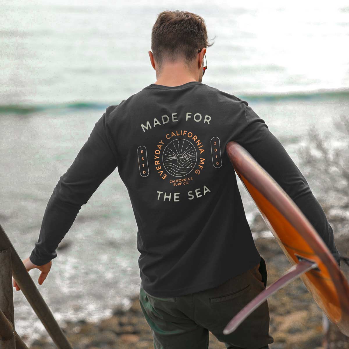 A model carries a surfboard while wearing an "Everyday California" "Made for the Sea" long sleeve