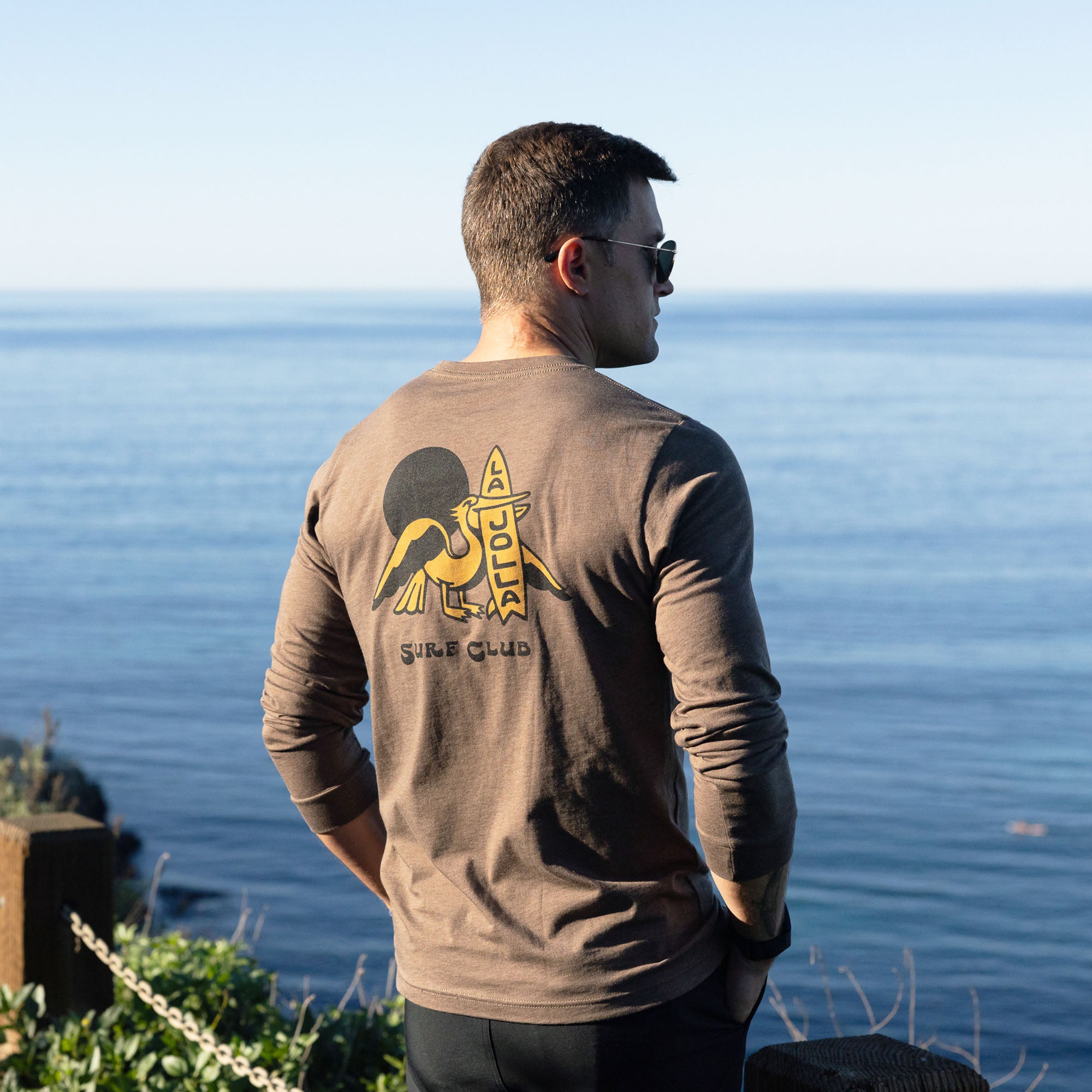 Man wearing a brown long-sleeve shirt with a logo, standing by the ocean.
