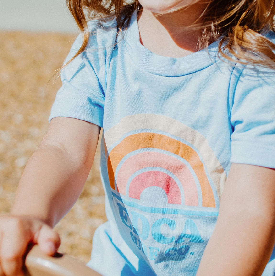 Lifestyle photo of a light blue shirt with a multi colored pastel rainbow on it, with text that reads EDCA SURF CO. beneath the rainbow.