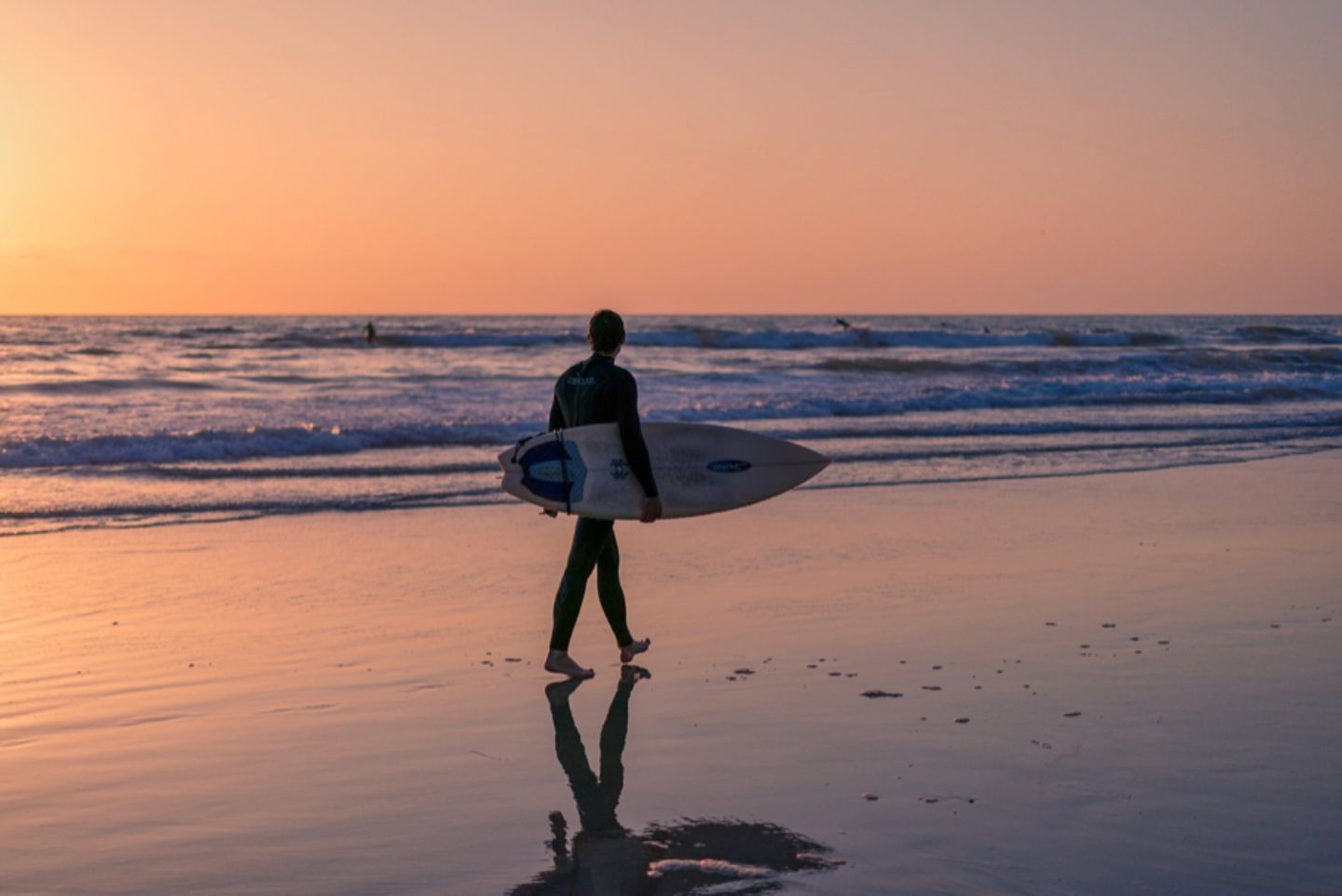 Sunset time and surfer holding surfboard on the beaches of San Diego. The water is calm and the sky is reflecting off the water and sand. 