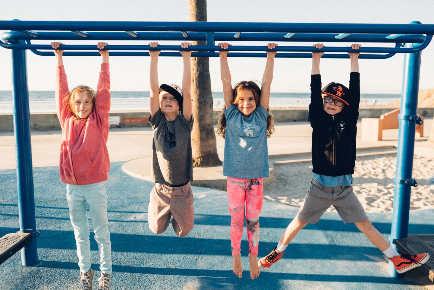 Multiple kids hanging from the monkey bars on the beach two girls and two boys wearing everyday California clothes