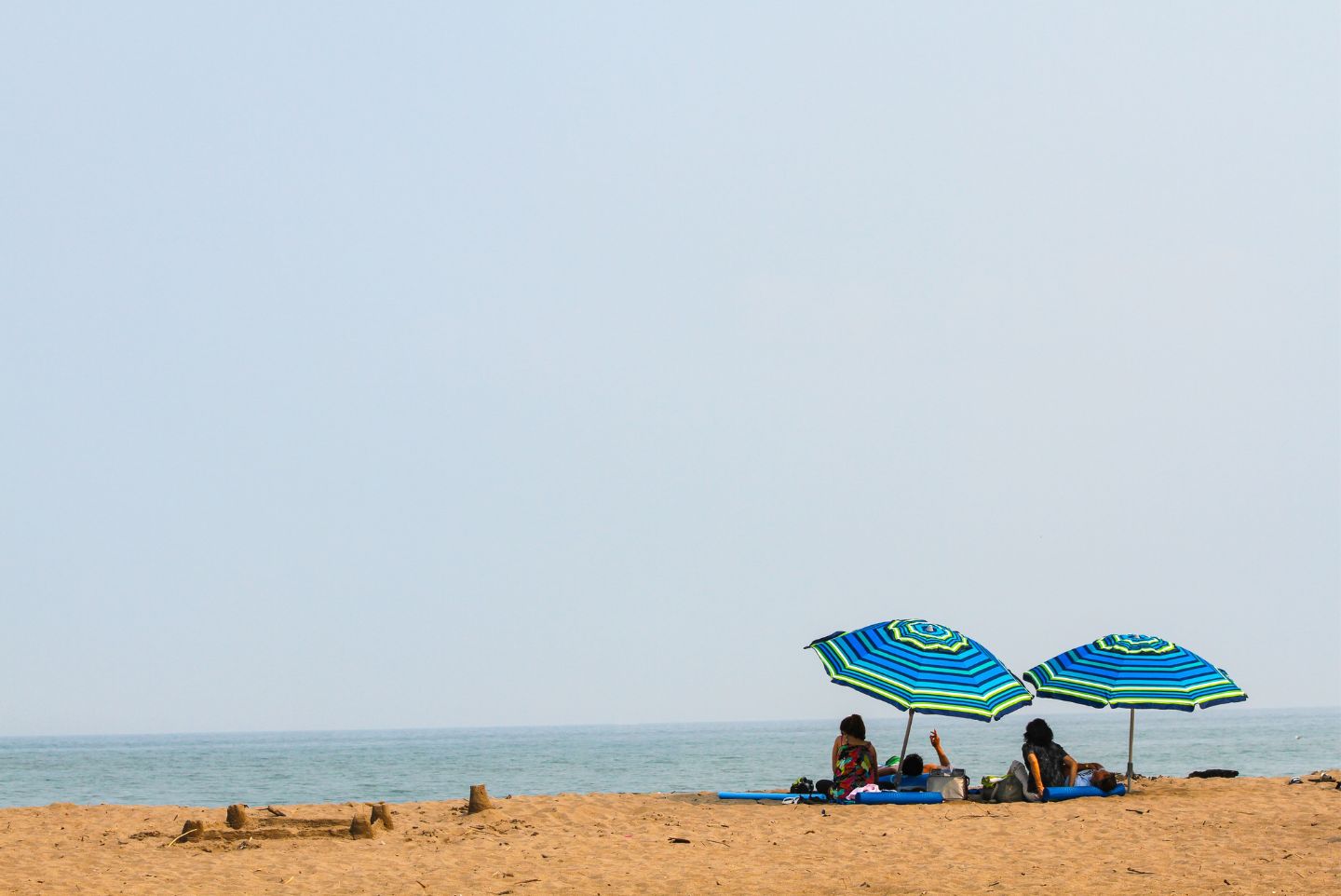 two people sitting on a sunny beach with two blue umbrellas for sun protection