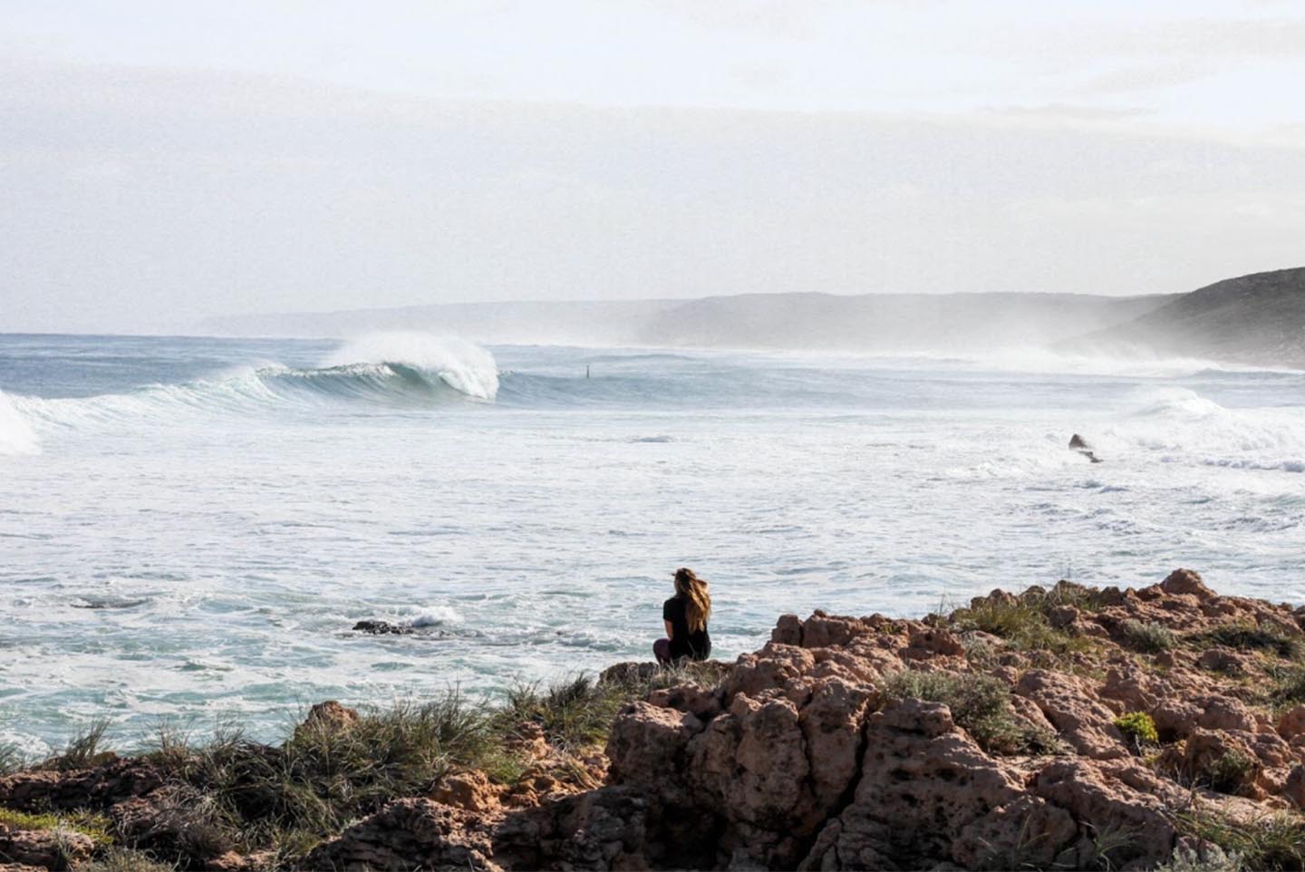 A girl sitting on a rock in front of the ocean with her back turned to the viewer. The sky is gray and there is a wave breaking in the distance in California.