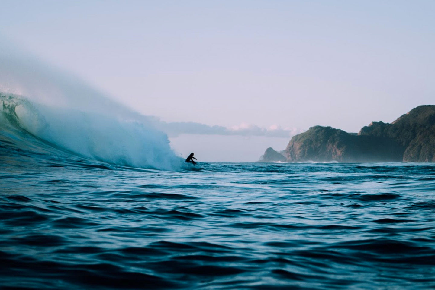 Surfer riding a wave in the ocean, with sea cliffs in the background. The surfer is the only person in the water.