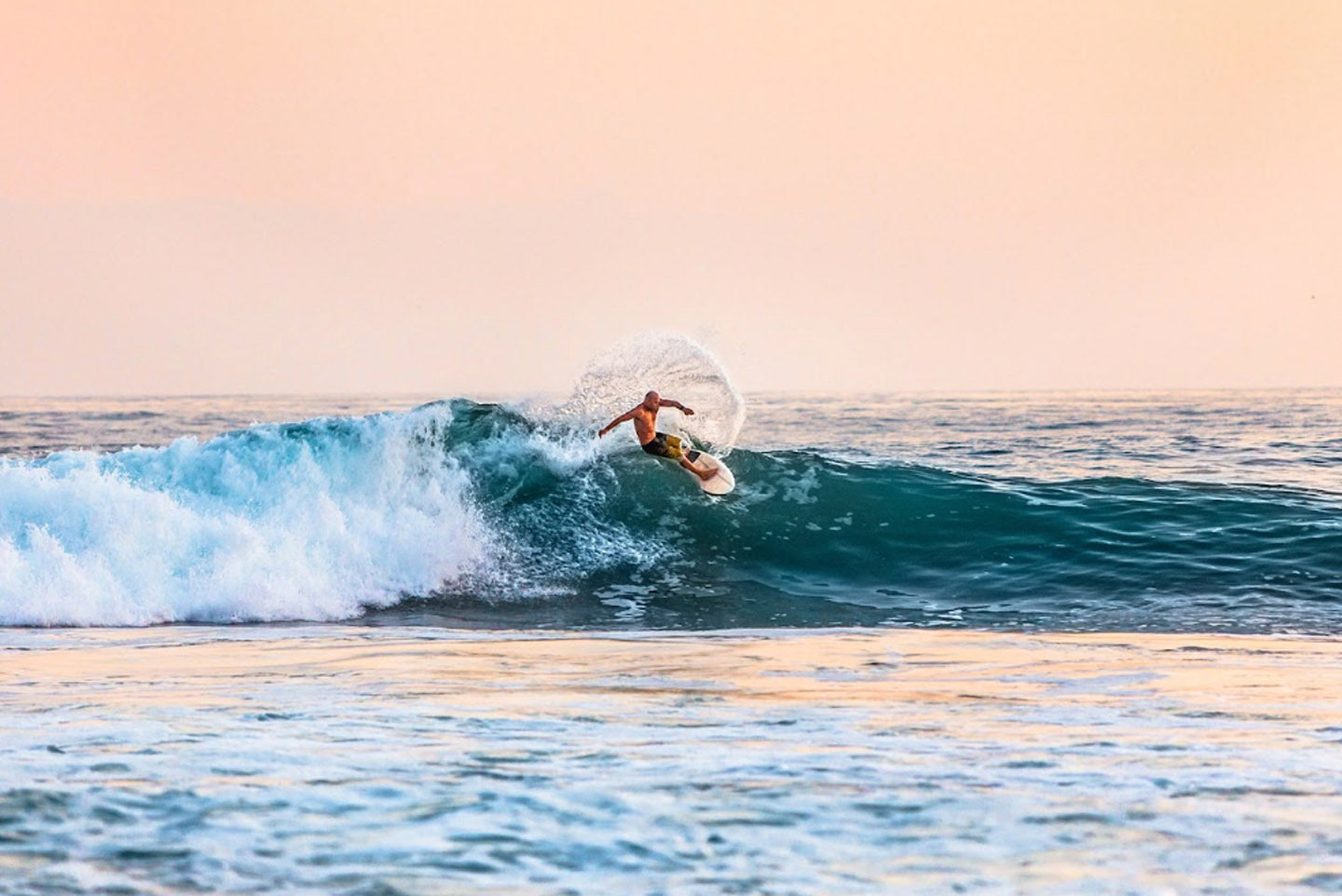 Surfer in the ocean riding a wave. There is nobody else in the water, and the sun is setting in the distance.