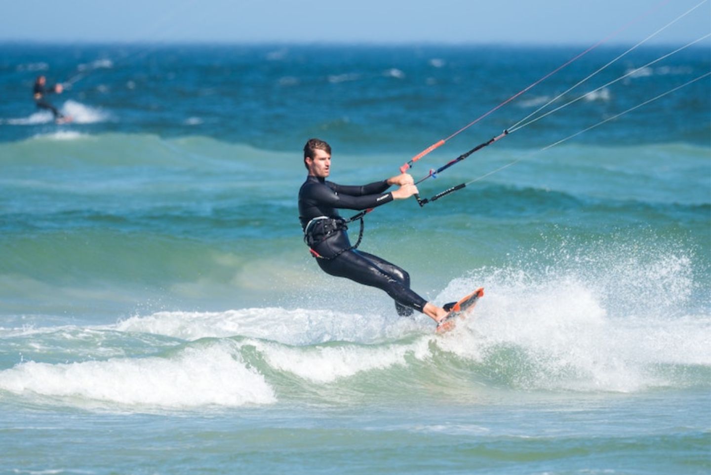 Man in a full wetsuit wind surfing holding onto his sail and surfing the blue water with blue skies. 