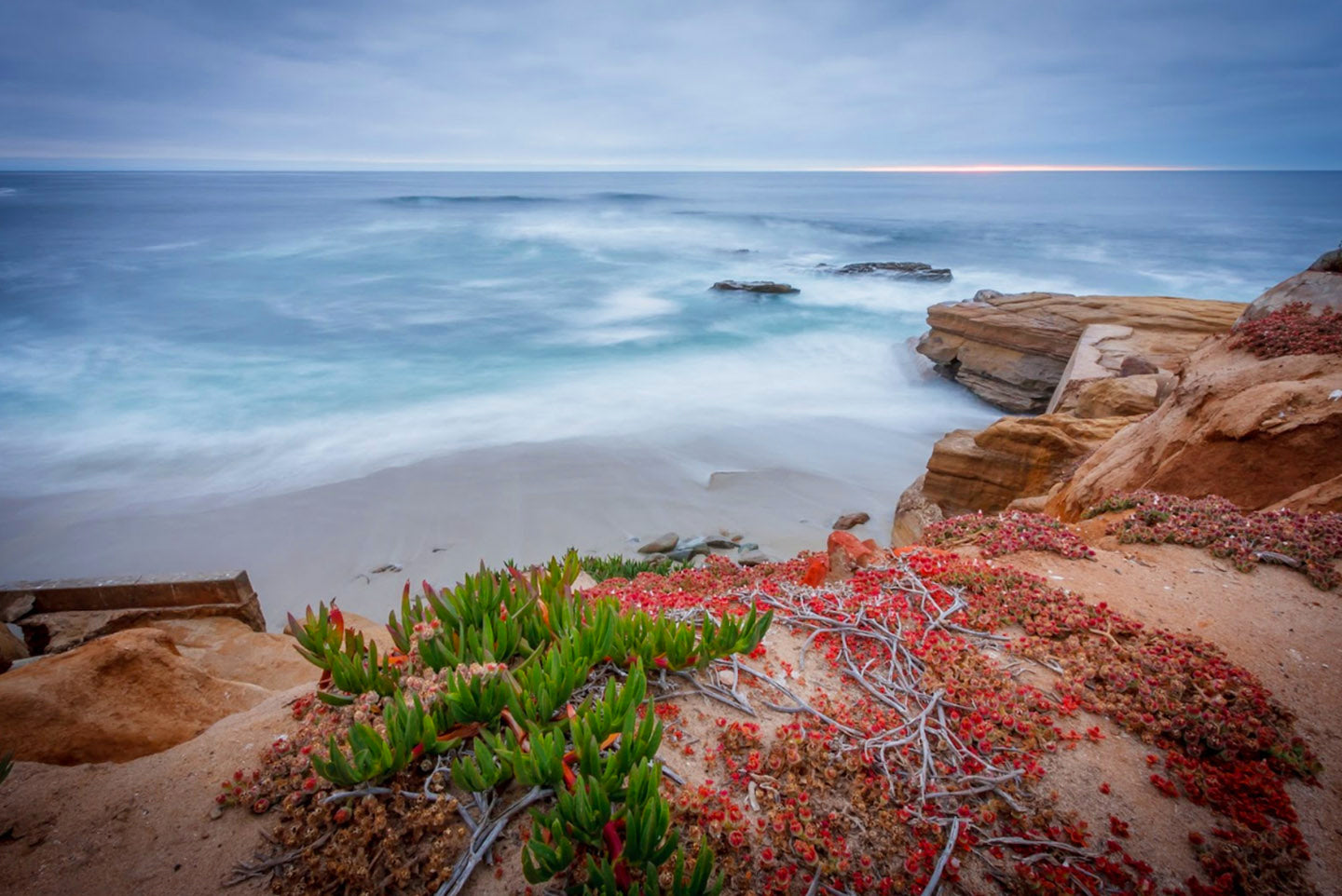 A photo of La Jolla Shores beach from the side of a cliff. The sky is overcast and gray.