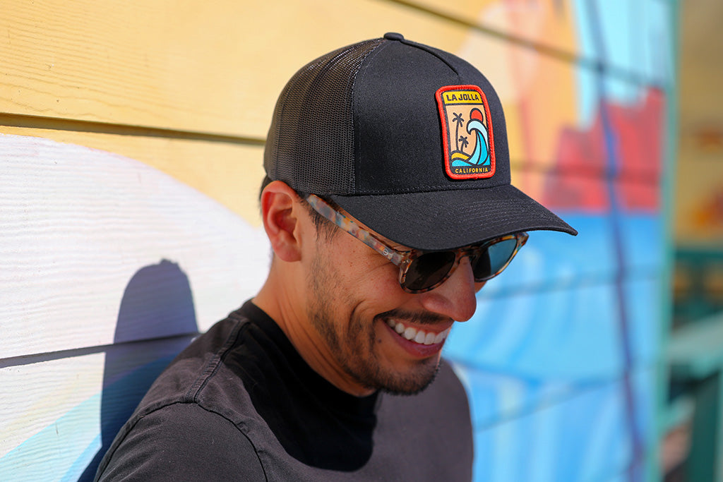 Photo of a man smiling, wearing sunglasses and a black hat with a patch on it that says "La Jolla - California". Everyday California Headwear.
