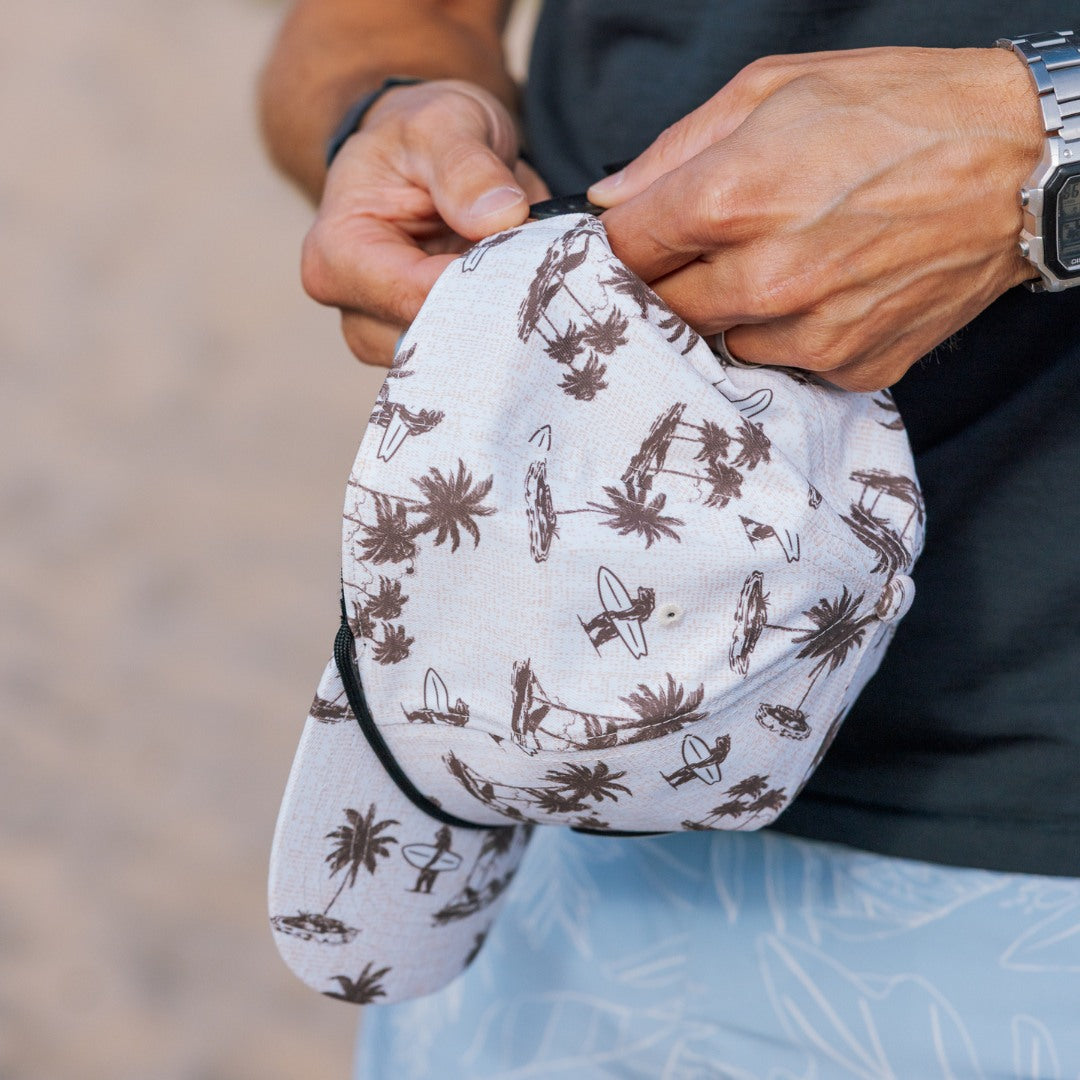 Person holding a white cap with brown patterns, Coastal Division Hat, wearing a black shirt and blue shorts.