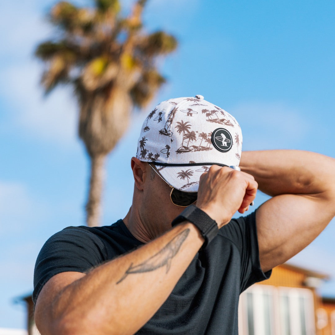 Man wearing the Coastal Division Hat with palm tree design against a blue sky and palm tree background