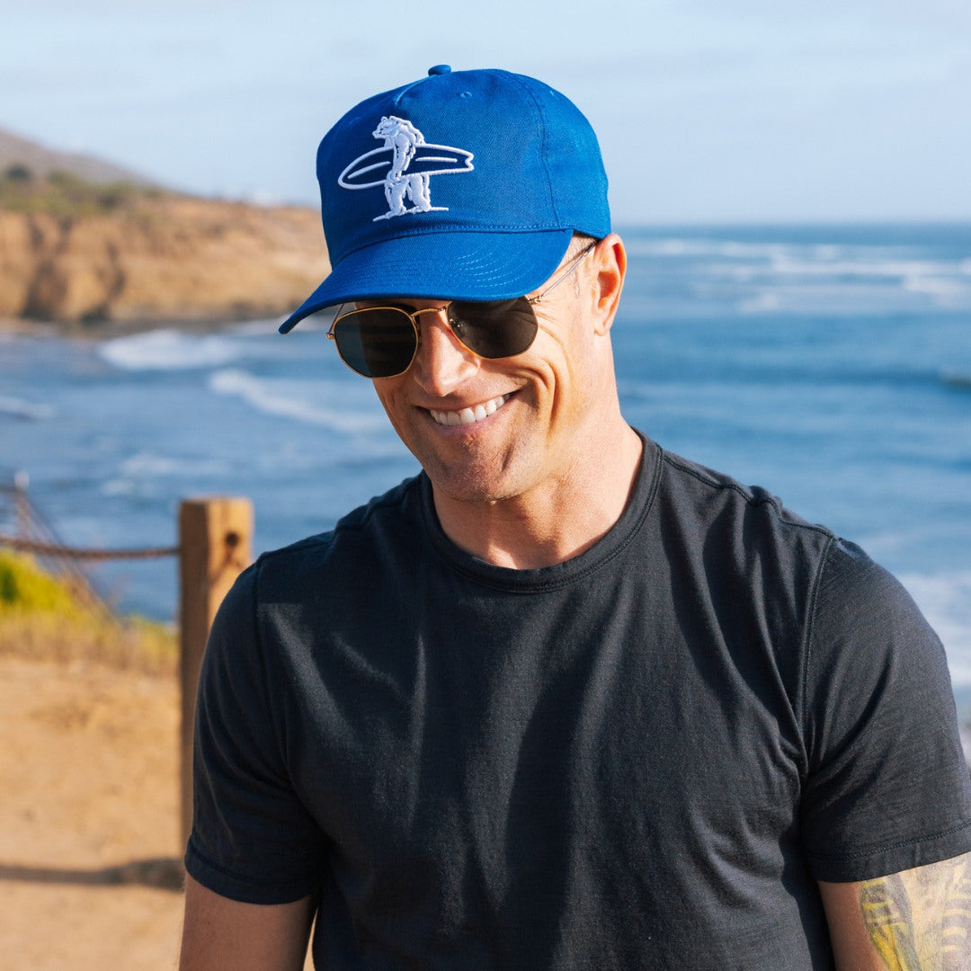 Man wearing a blue cap with Everyday California logo and sunglasses with a scenic ocean background