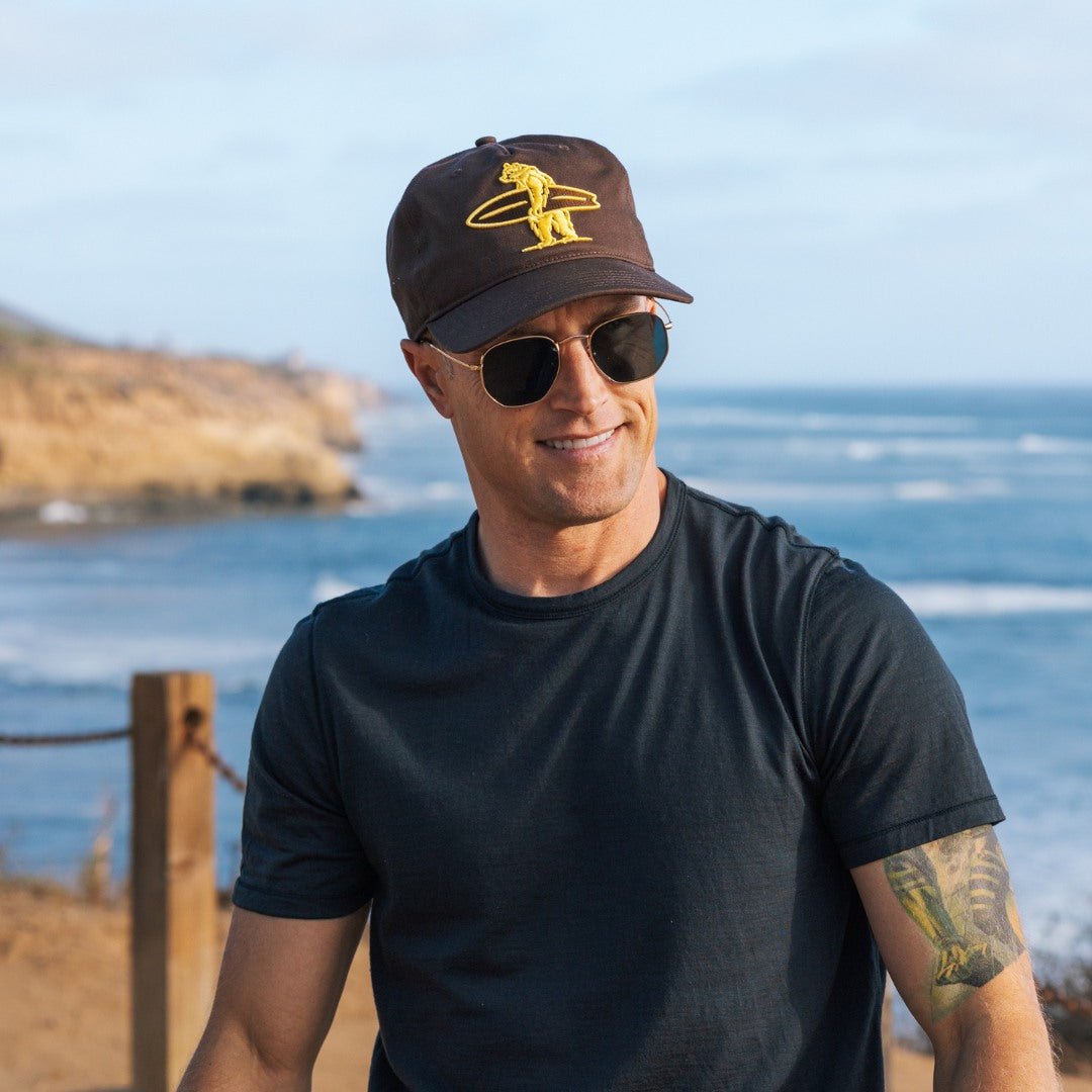 Man wearing a brown cap with Everyday California logo and sunglasses with a scenic ocean background