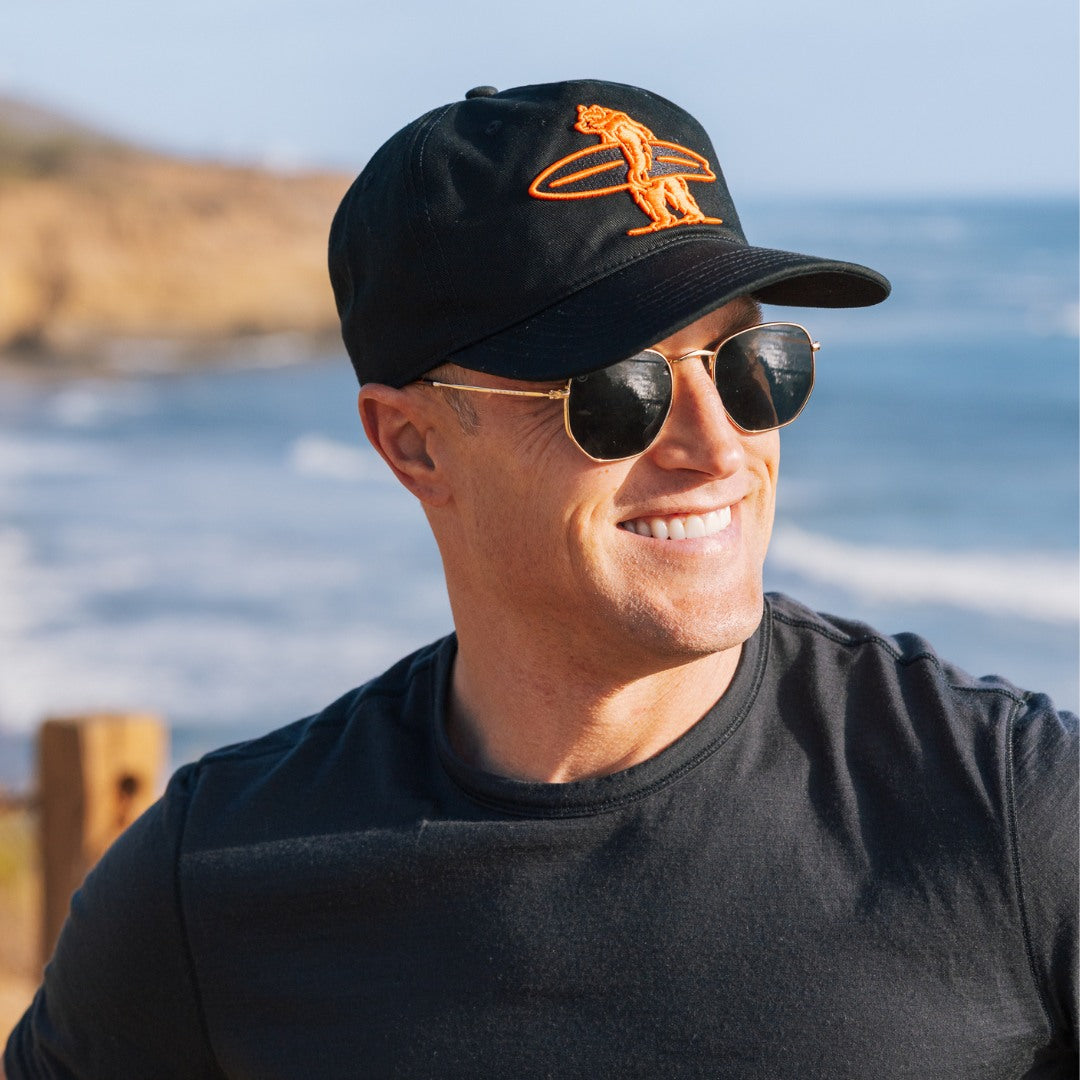 Man wearing a black cap with an orange Everyday California logo and sunglasses, standing by the ocean.