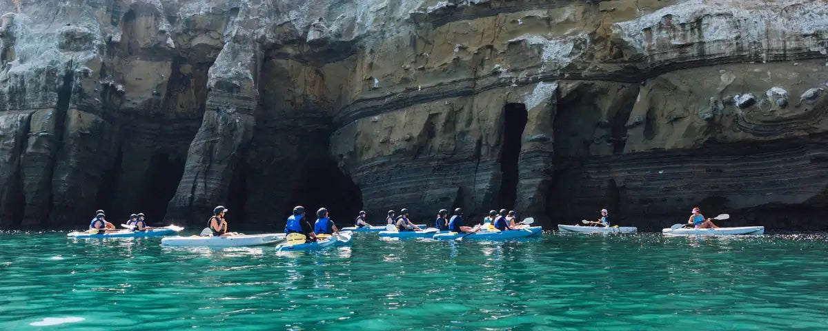 A large group of around 15 kayakers in the ocean, in front of the entrance of the Seven Sea Caves in La Jolla.