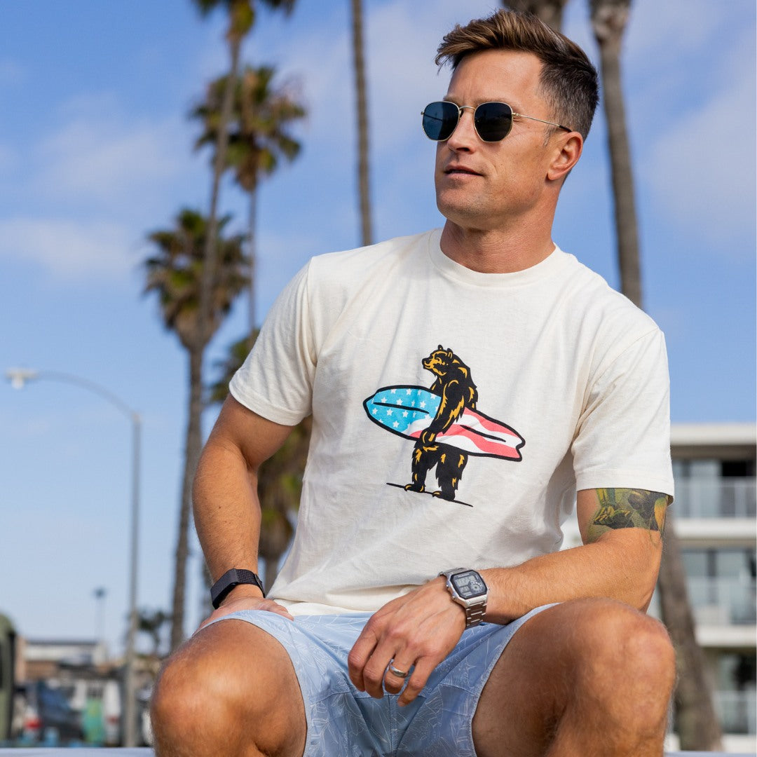 Man wearing a white t-shirt with a bear holding a surfboard and American flag design, sitting outdoors with palm trees and blue sky in the background.